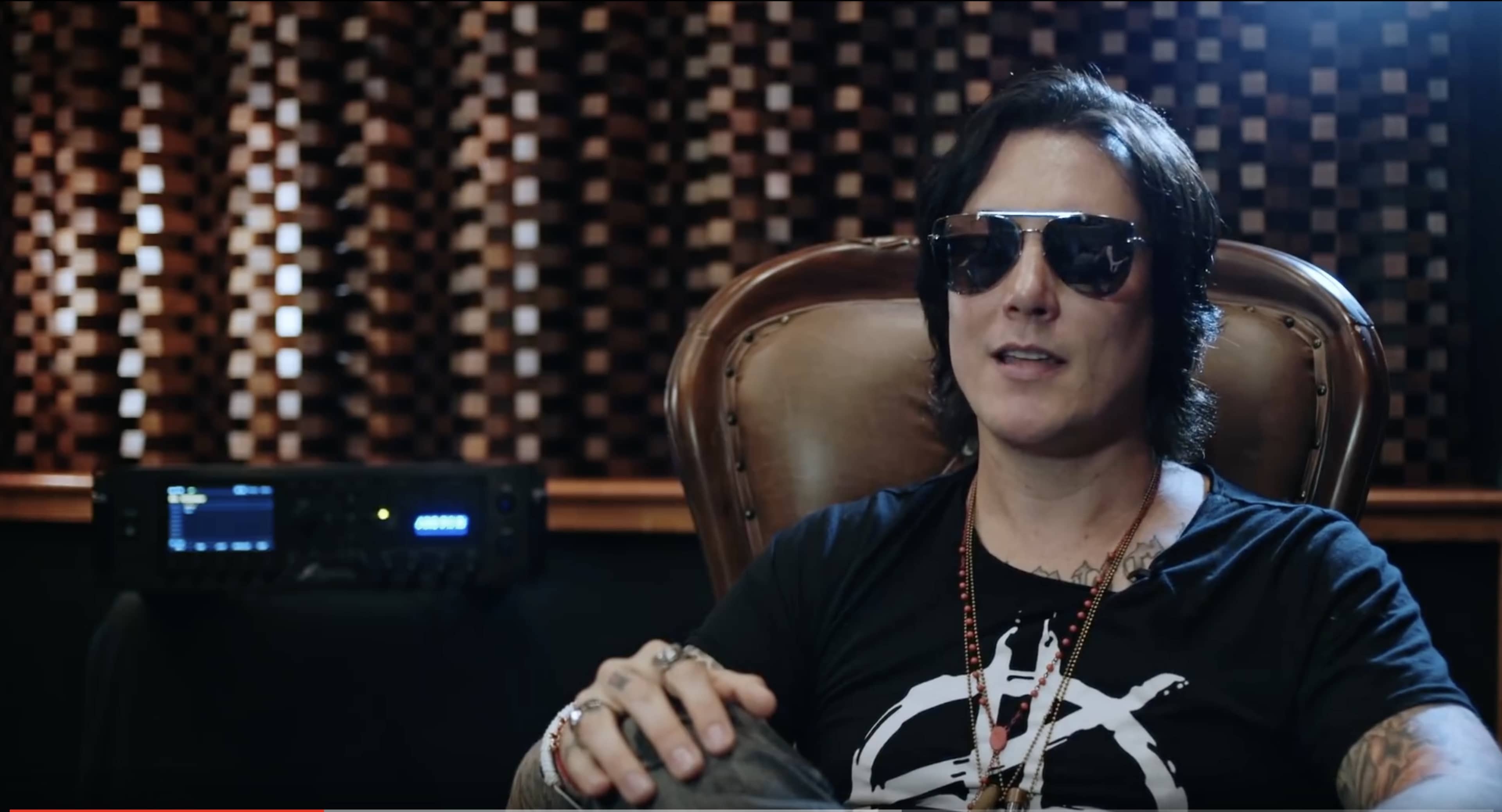 A musician wearing sunglasses sits in a chair in a studio with a patterned wall and recording equipment in the background.