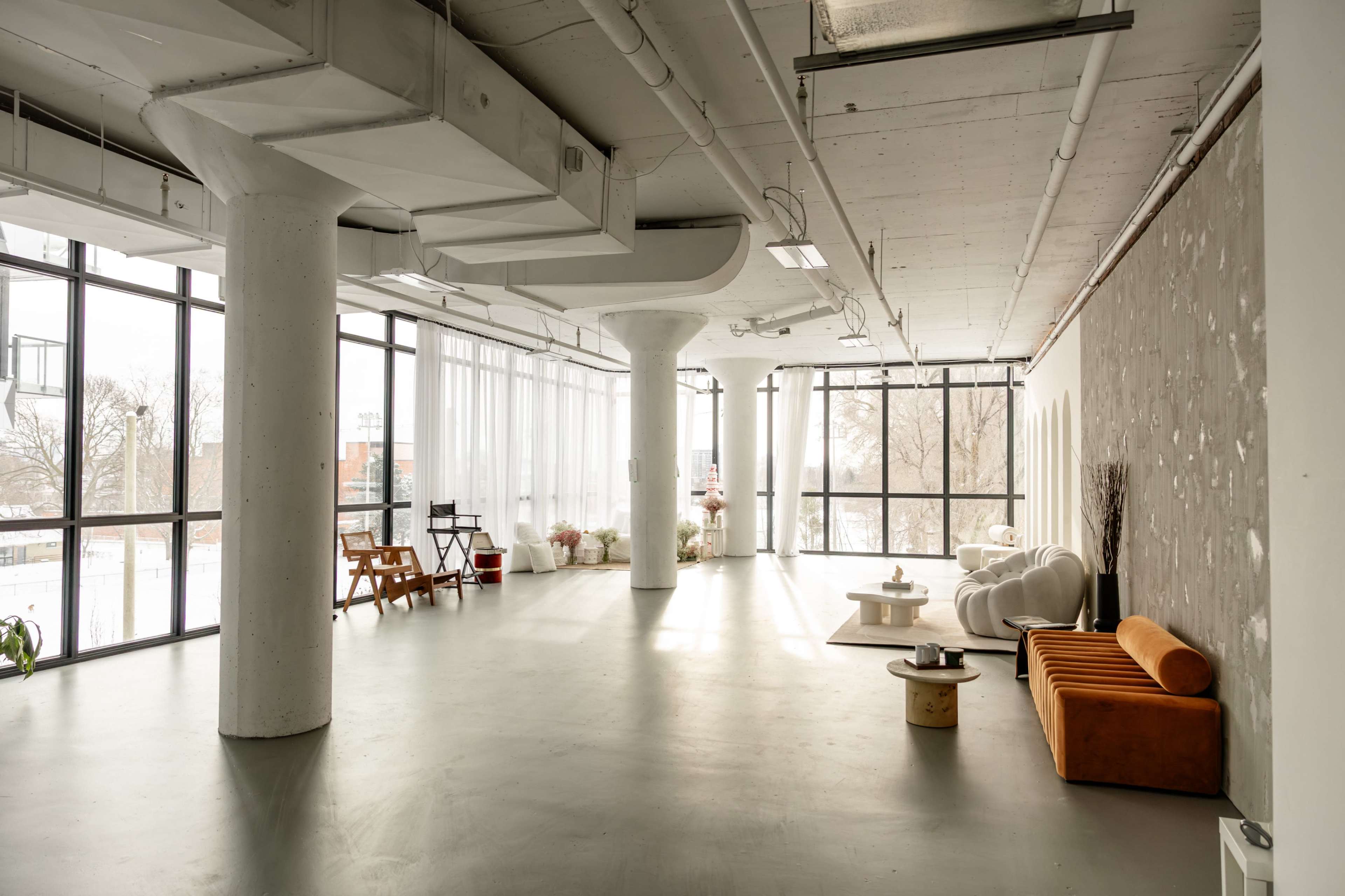 A spacious, modern interior featuring large windows, concrete pillars, and a mix of seating, including a sofa and tables.