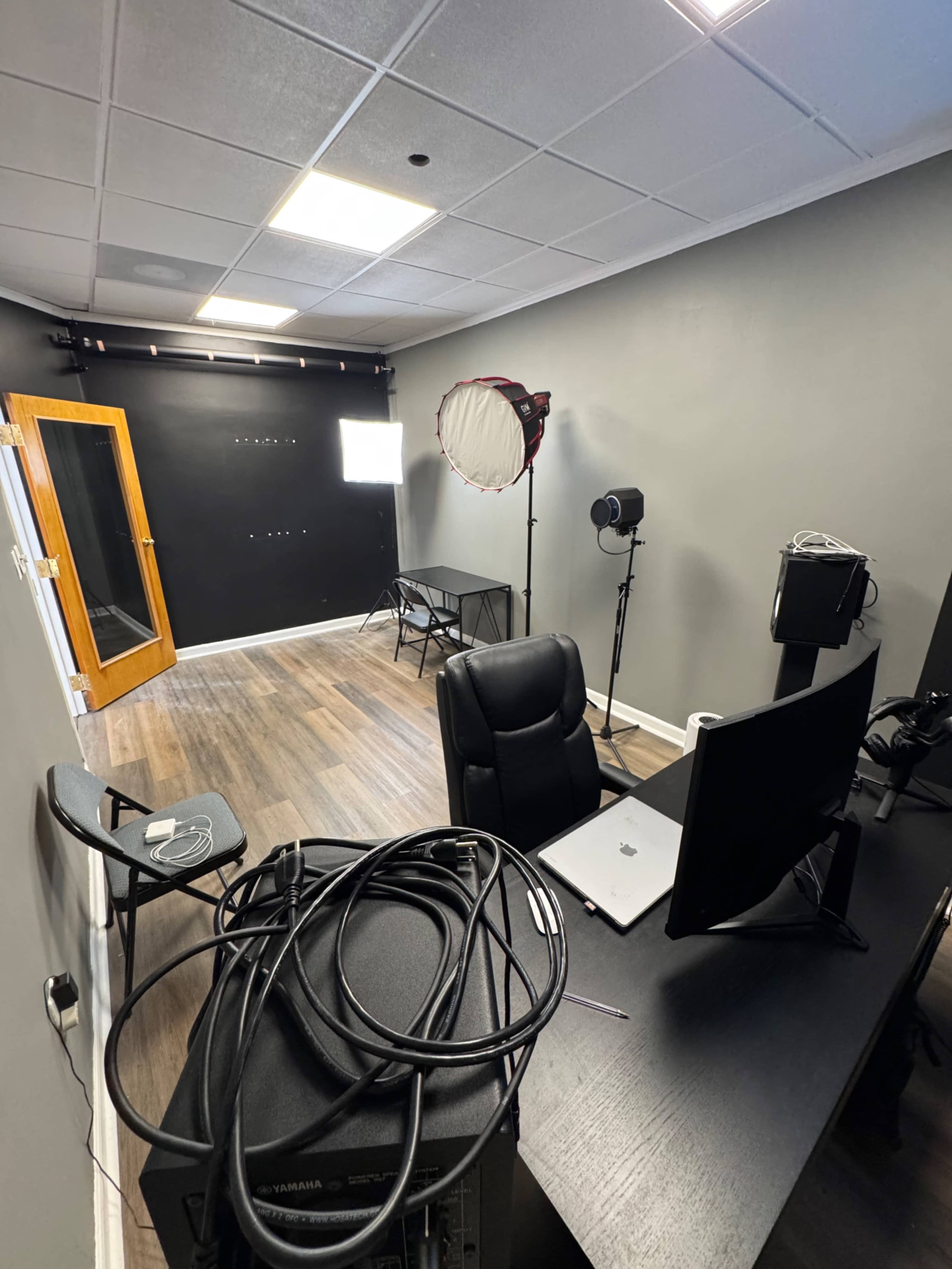 The image shows a small studio with a black wall, a lighting setup, a desk with a monitor, and various cables on the floor.