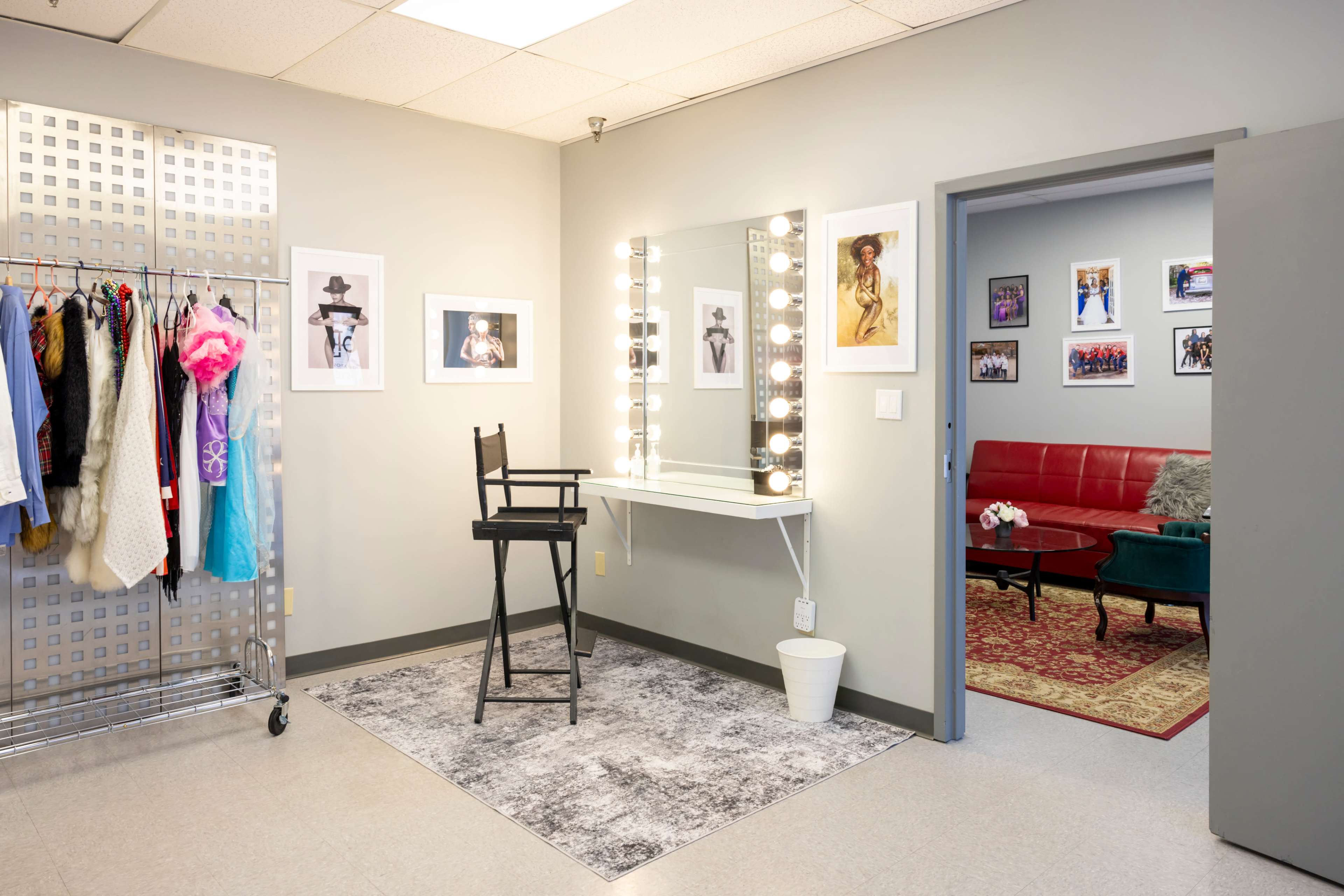 The image shows a well-lit dressing room with a mirror surrounded by lights, a chair, a clothing rack with various costumes, and a doorway leading to a red seating area decorated with framed photographs.