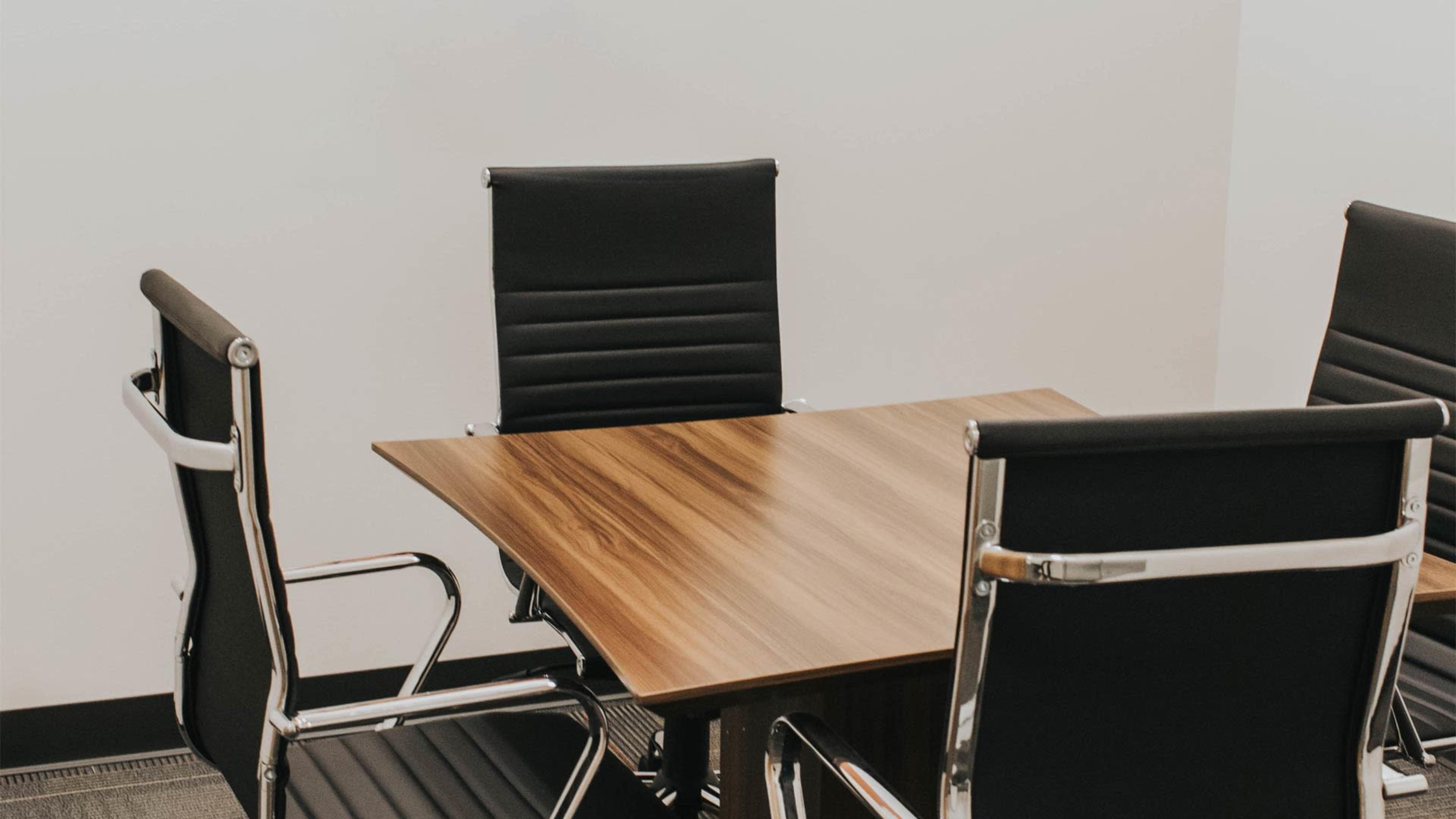 A wooden conference table with four black leather chairs is set against a plain white wall.