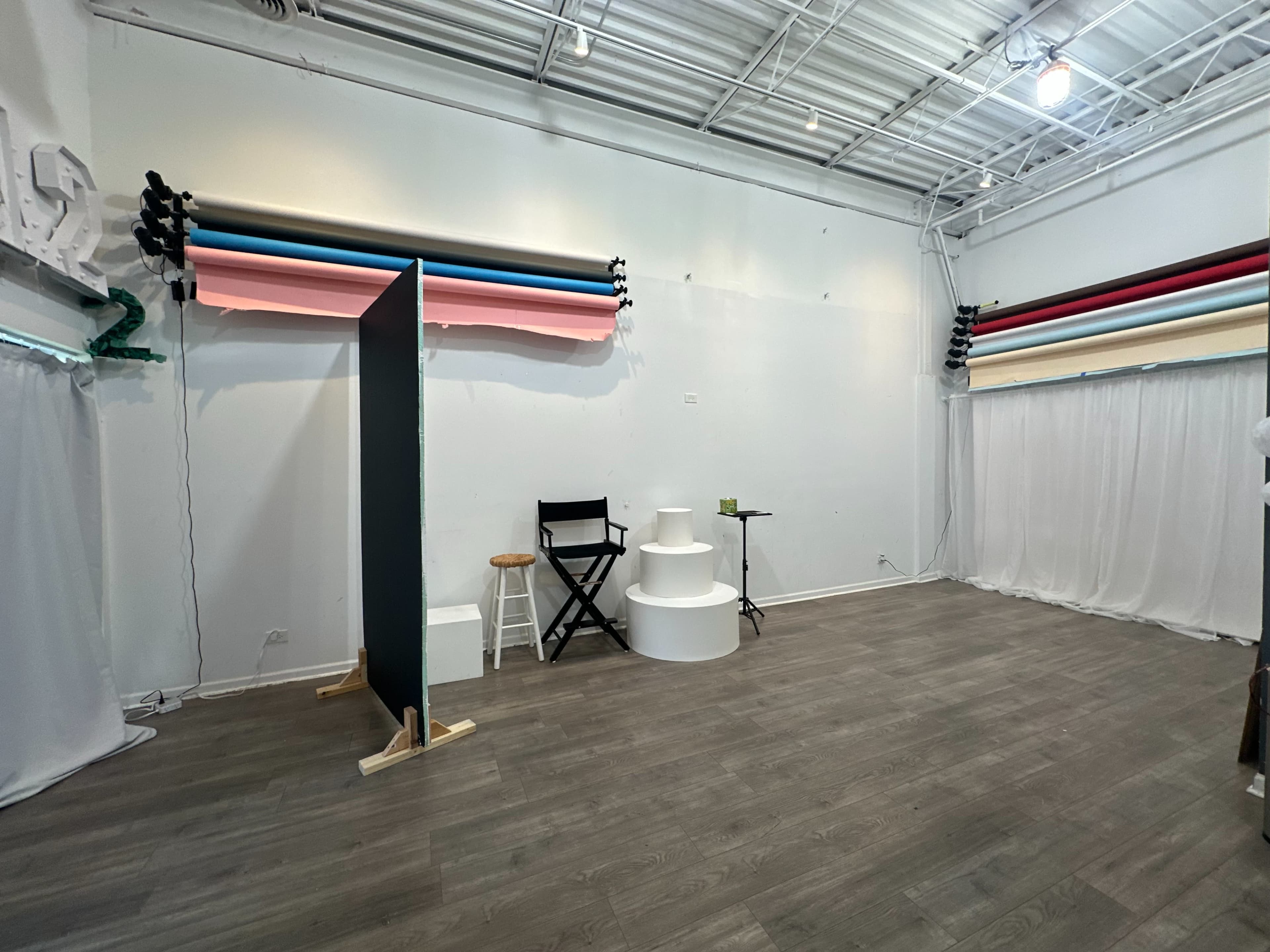 The image shows a spacious studio with a large, empty floor, multiple rolls of backdrop paper displayed on the wall, a white circular pedestal, a chair, and a small table.