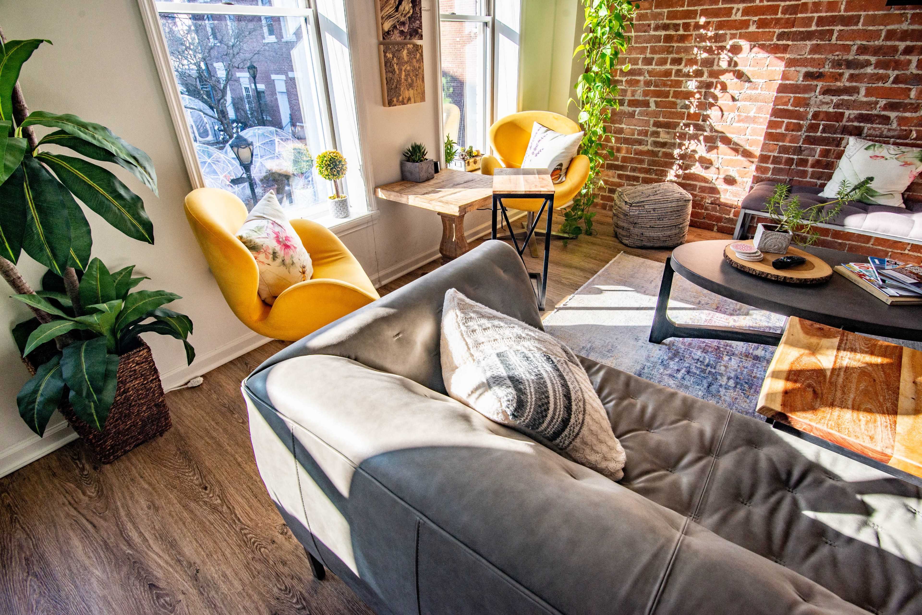 A cozy living room with a gray sectional sofa, two yellow armchairs, potted plants, and a brick wall, bathed in natural light from large windows.
