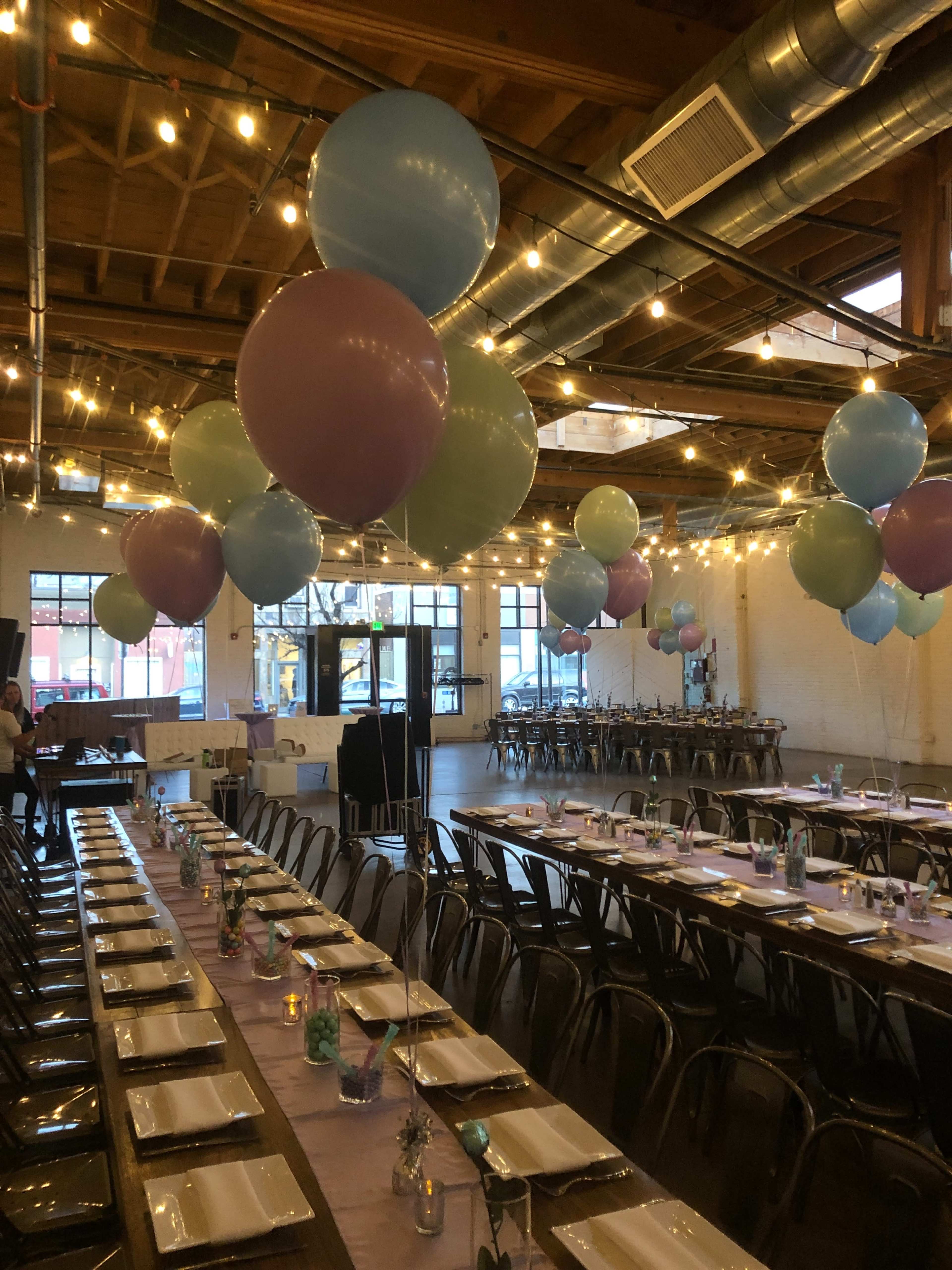 The image shows a spacious event venue decorated with large pastel balloons and string lights, featuring a long banquet table set with plates and glasses.