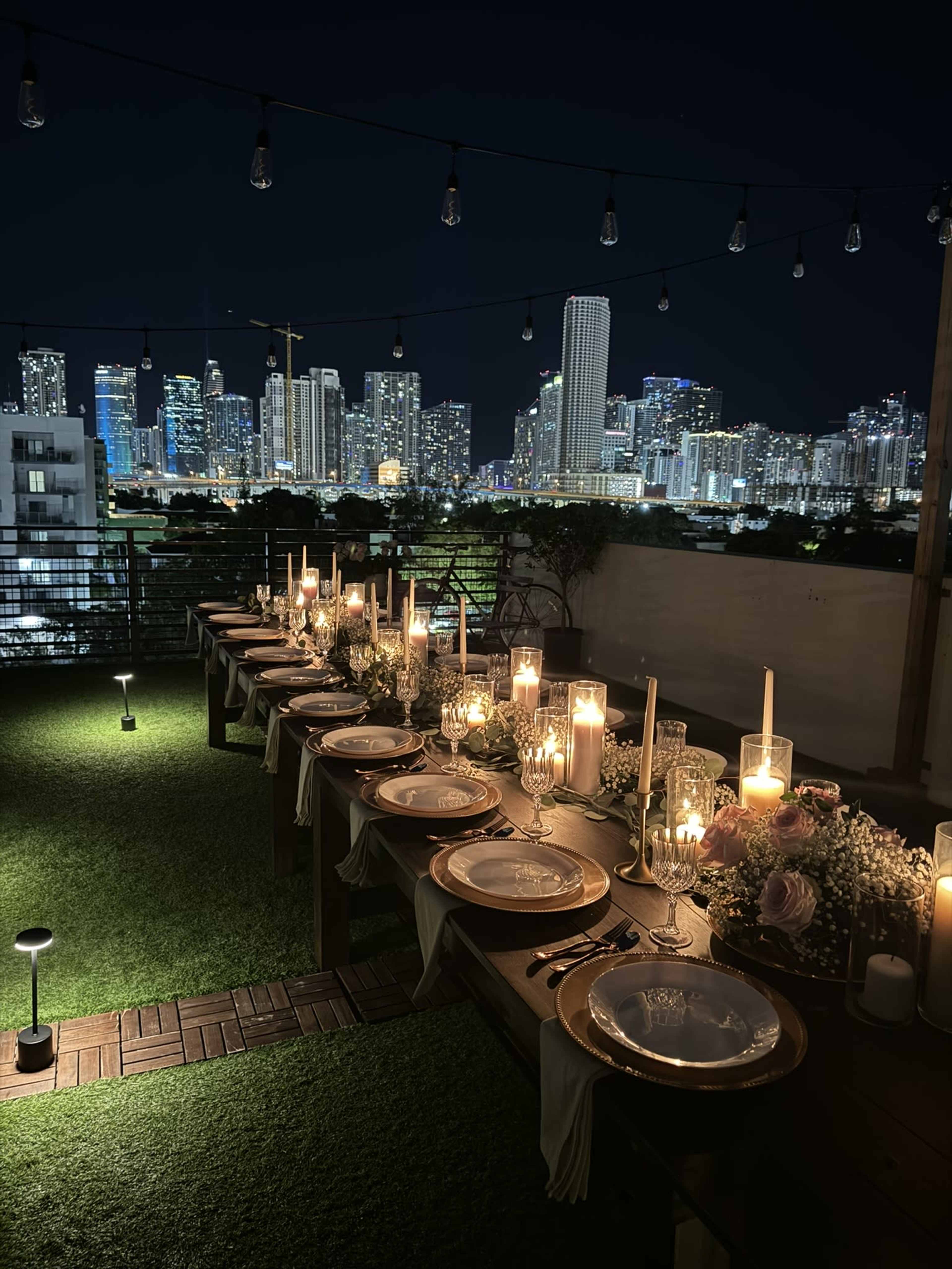 A long dining table elegantly set with candles and dinnerware overlooks a brightly lit city skyline at night.