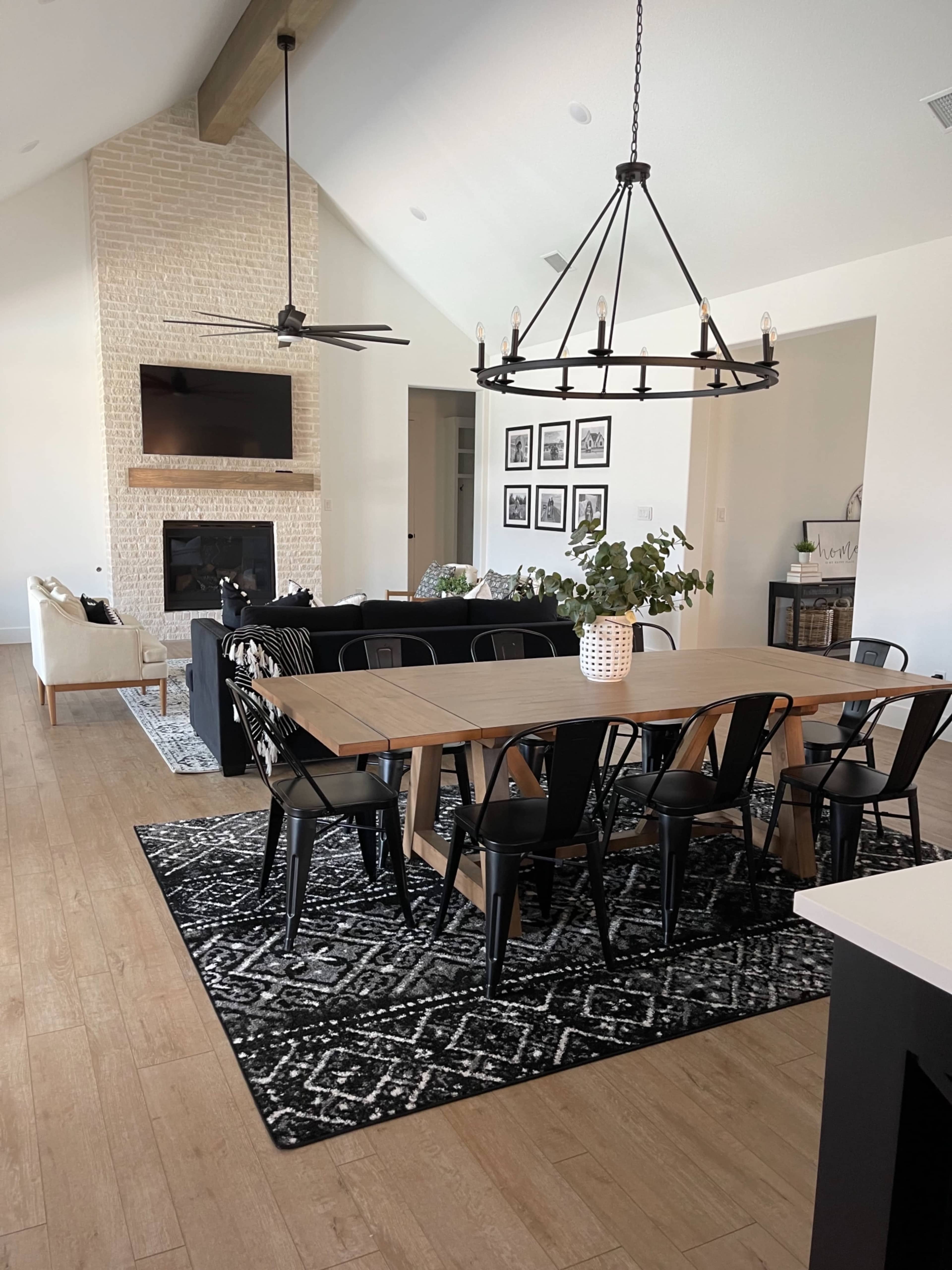 A modern dining area with a large wooden table surrounded by black chairs, set against a backdrop of a living room featuring a fireplace and wall-mounted TV.