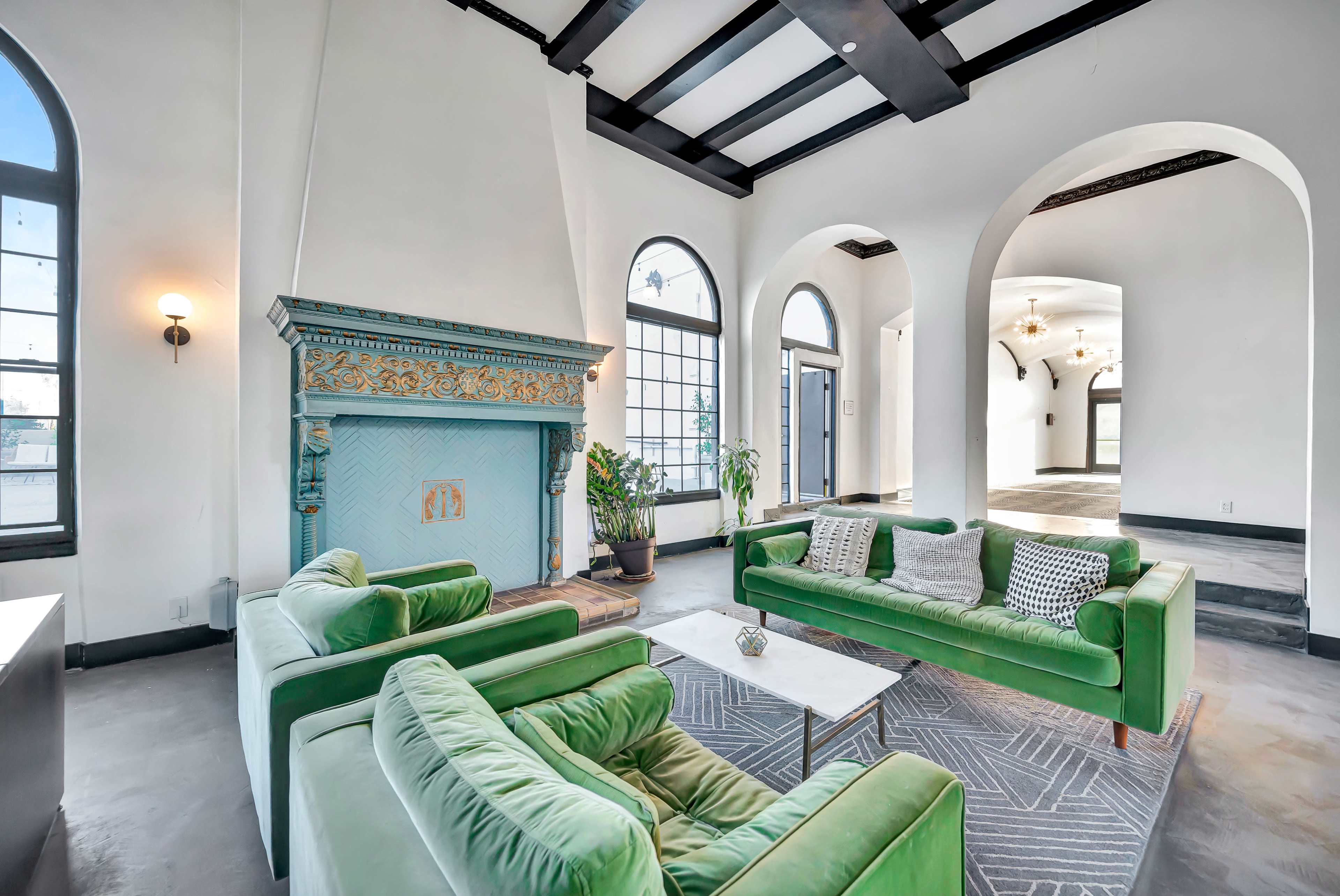 A spacious living area features green sofas positioned around a small table, with a decorative blue fireplace against a white wall and arched windows allowing natural light to enter.