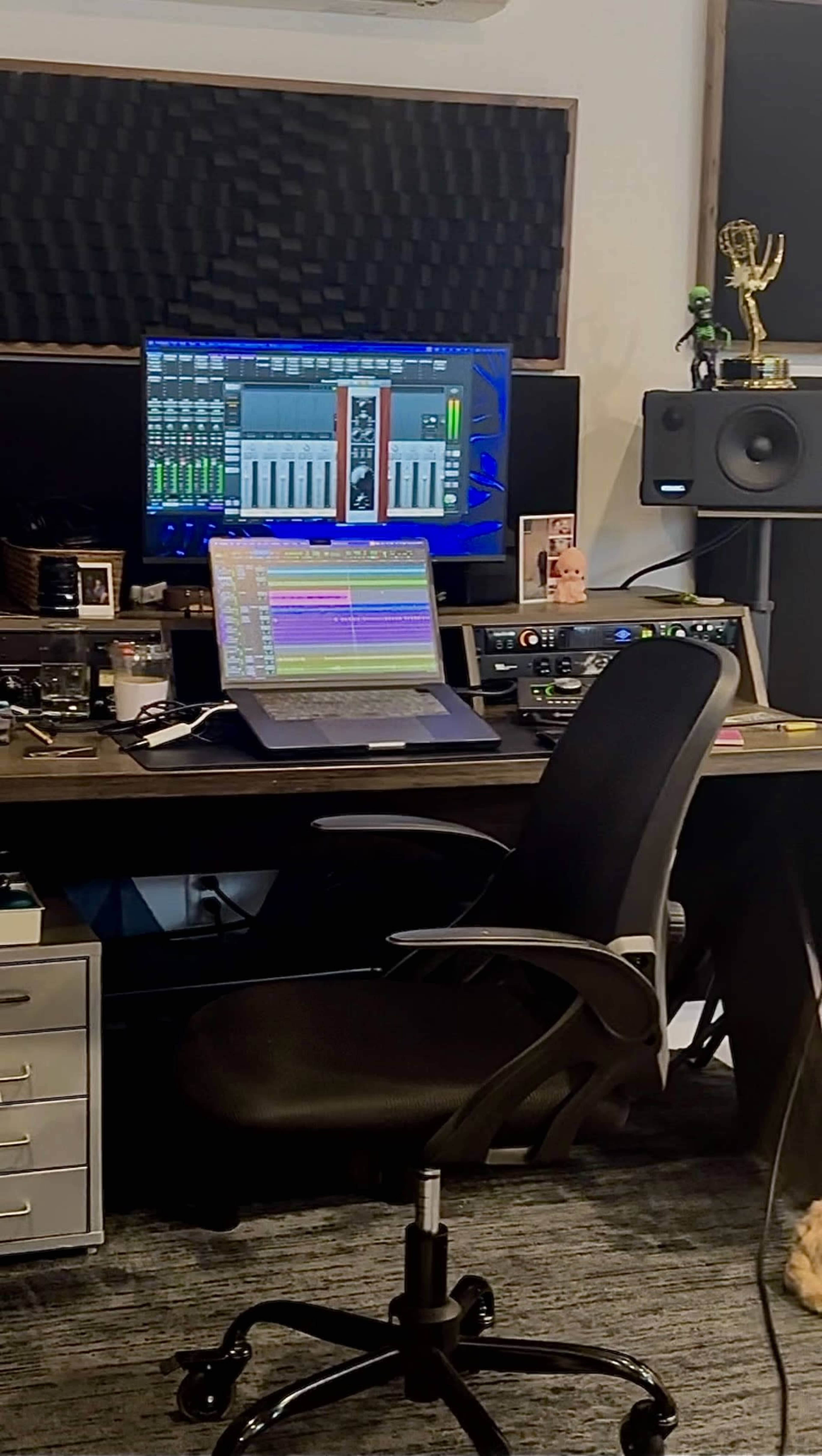 The image shows a music production studio setup with a computer, audio equipment, and a desk chair.