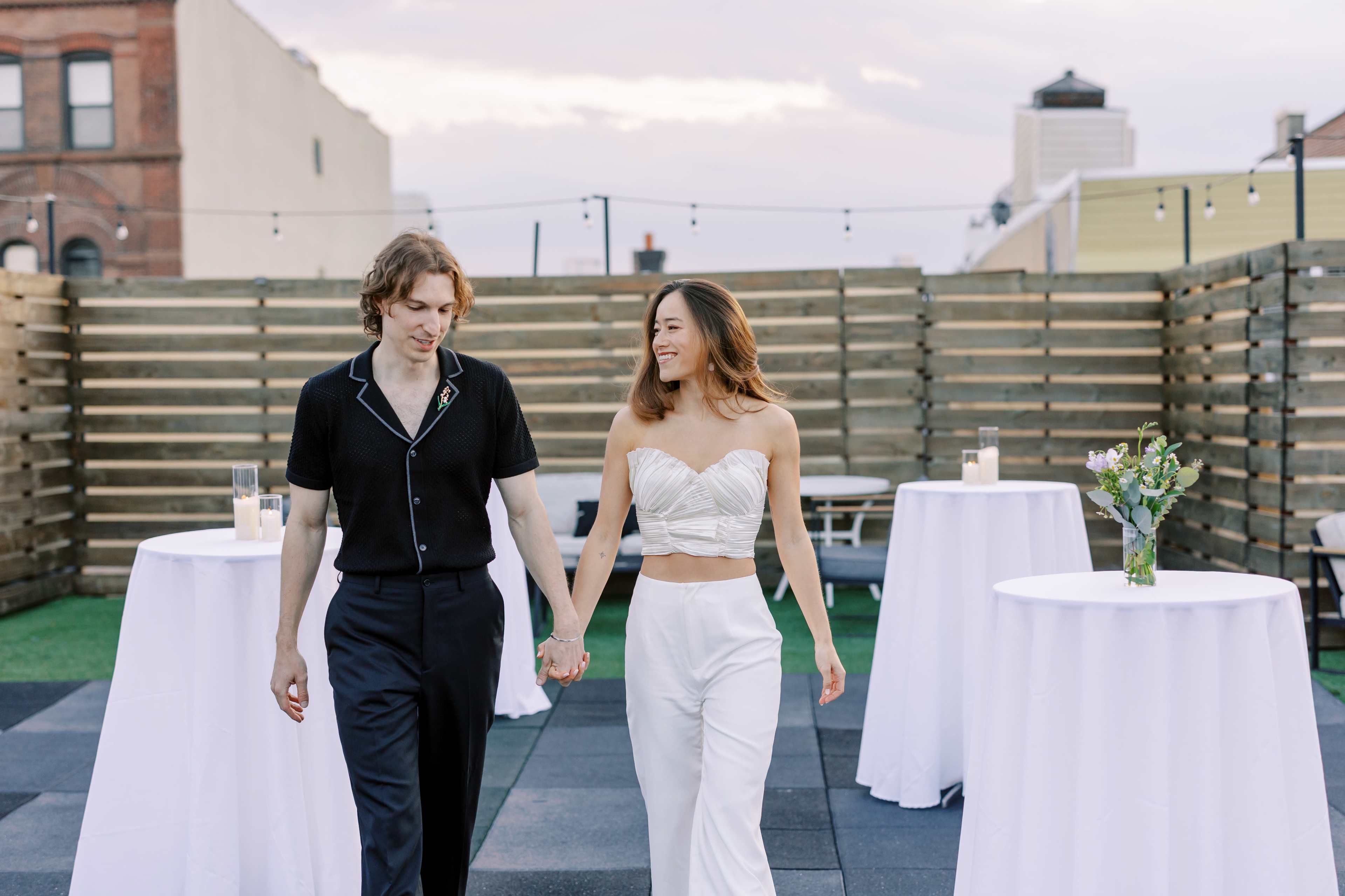 A couple walks hand in hand on a rooftop terrace adorned with white tables and soft lighting.