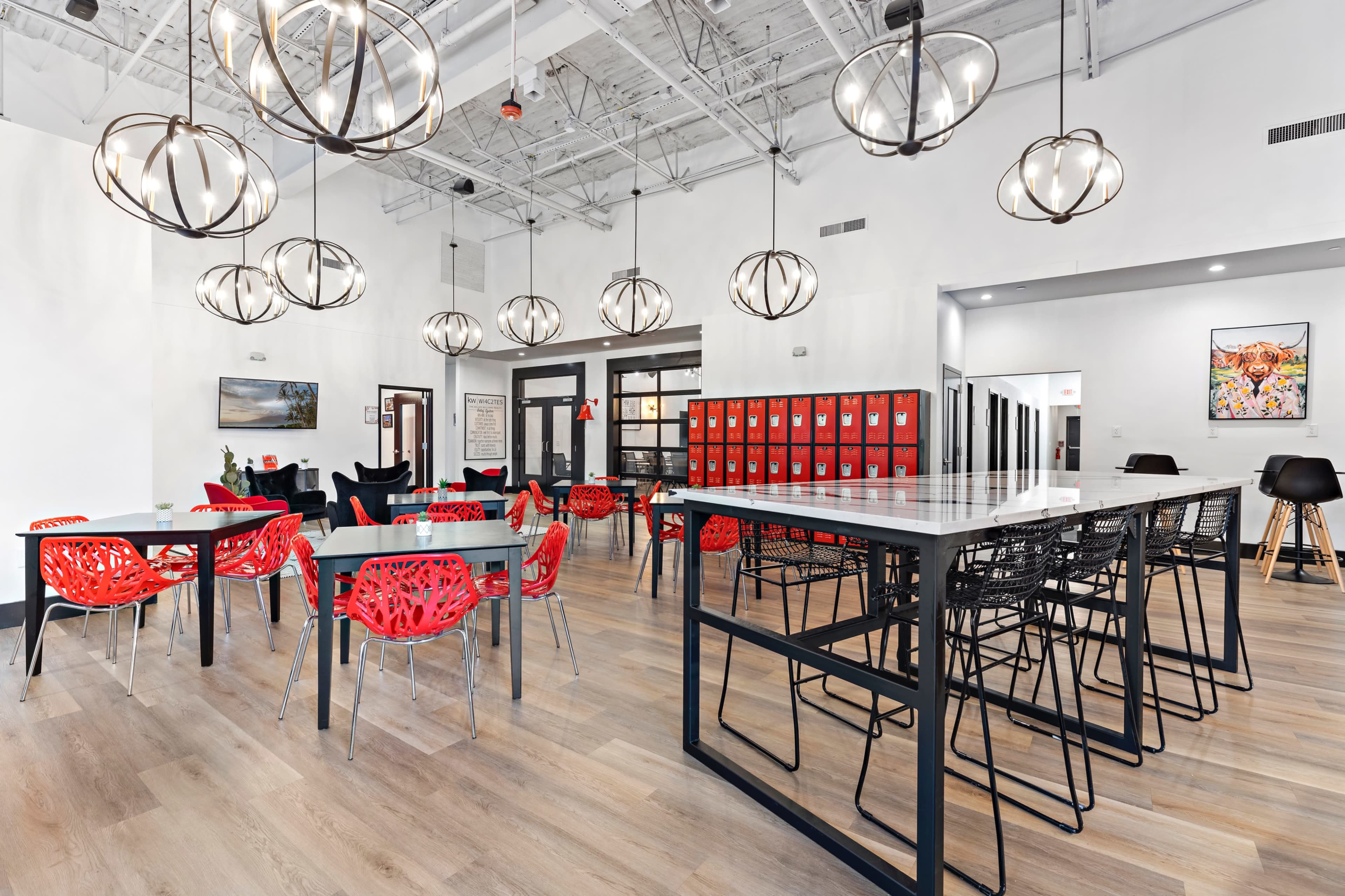 A spacious, modern communal area with tables and chairs, pendant lights, and red lockers in the background.