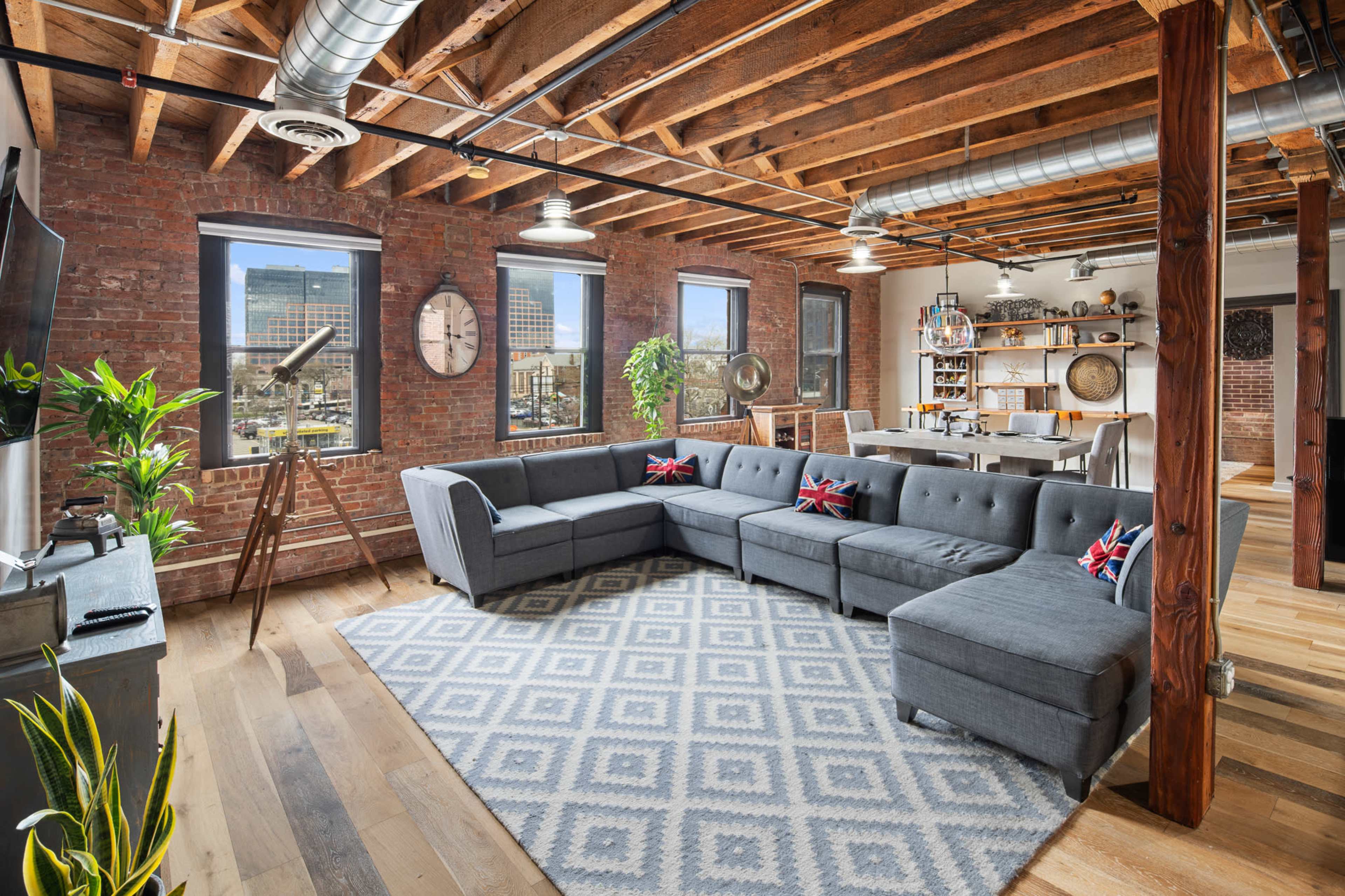 A spacious industrial-style living room with exposed brick walls, wooden beams, a large gray sectional sofa, and a dining area in the background.