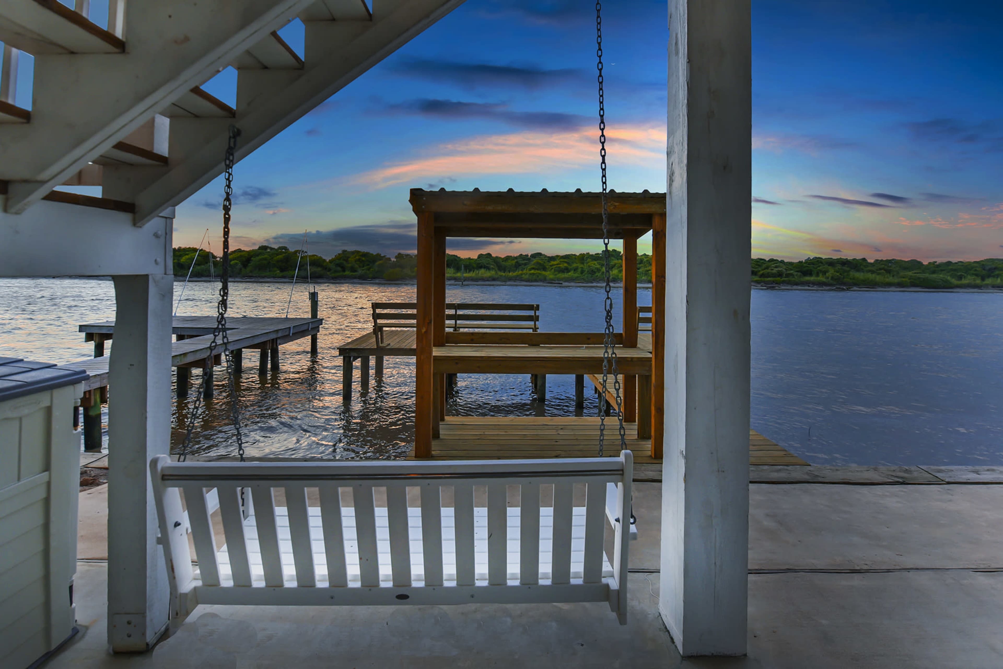 A white swing hangs under a staircase, overlooking a calm lake with wooden docks and a gazebo under a colorful sky.