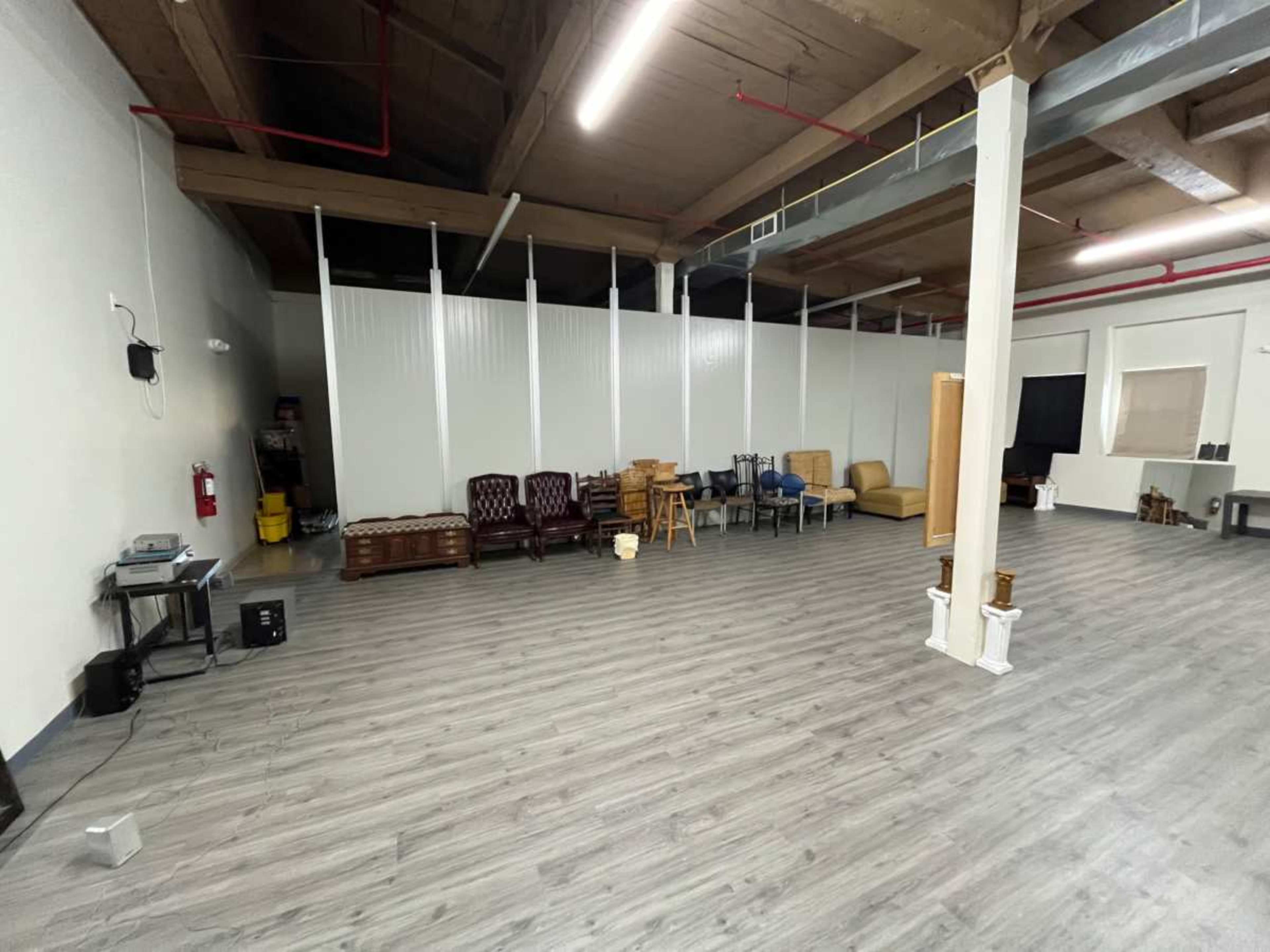 The image shows a spacious, empty room with wooden flooring and various pieces of furniture arranged against one wall.