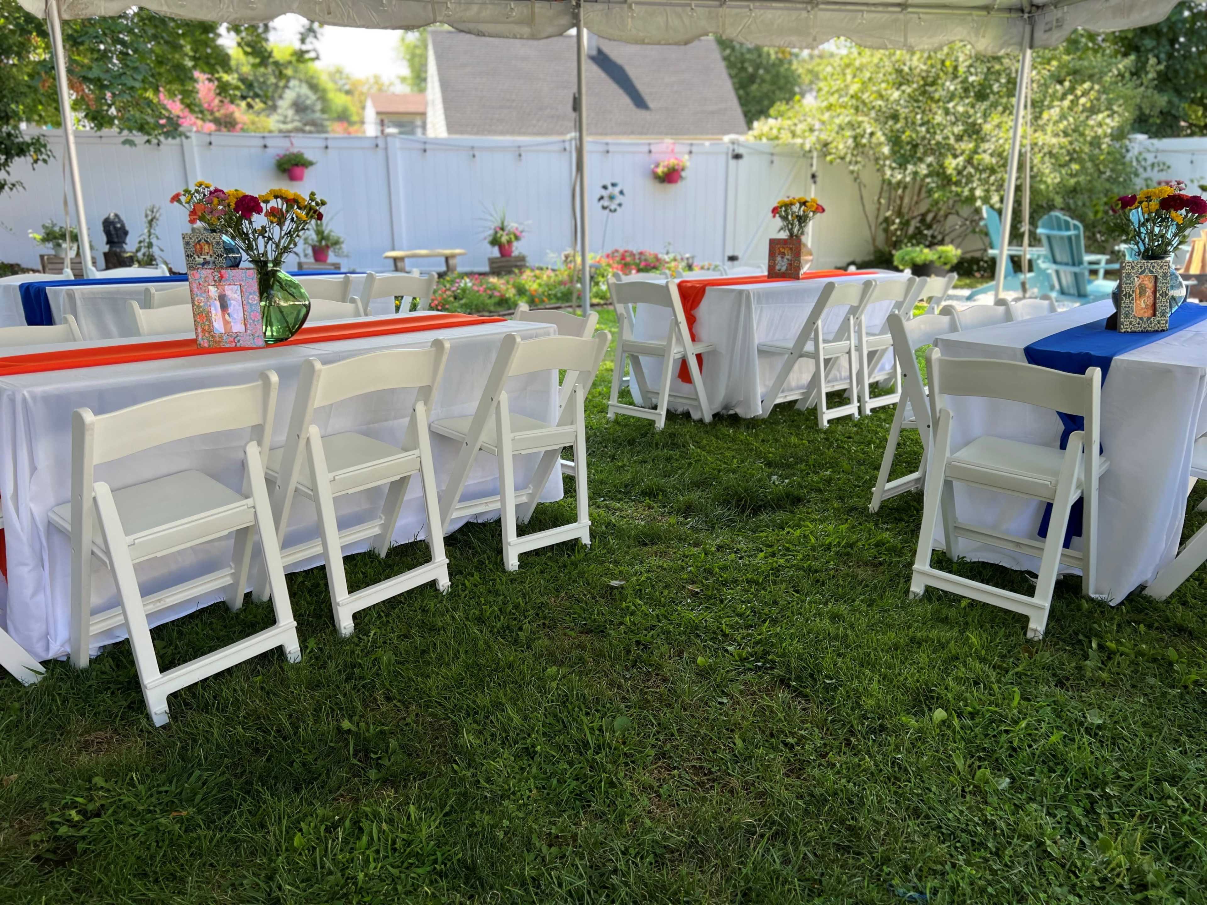 The scene shows a picnic area under a tent with decorated tables featuring flower arrangements and tablecloths in orange and blue, set up on a grassy lawn.