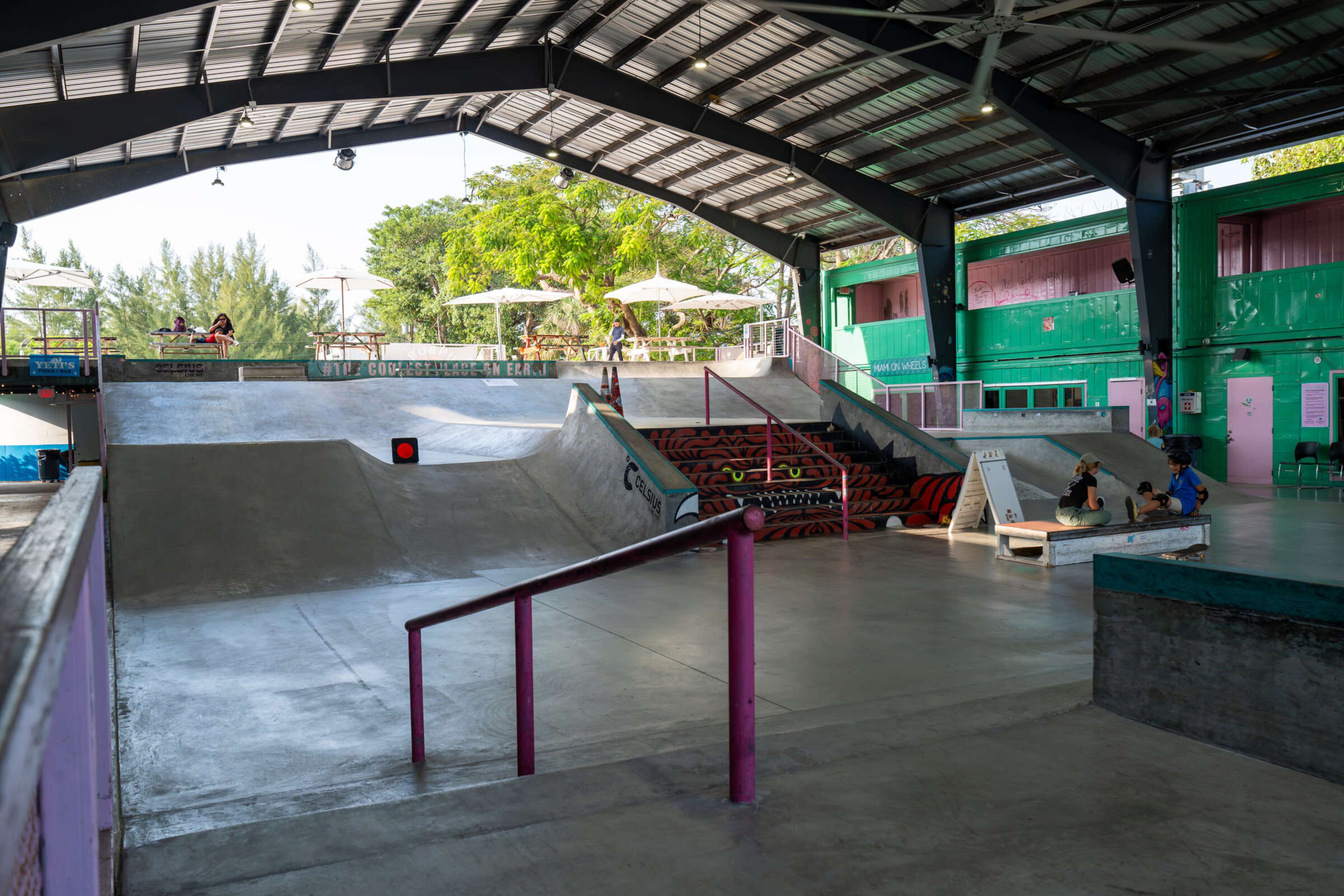 The image shows an indoor skate park featuring ramps, rails, and seating areas, with sunlight filtering through large windows.