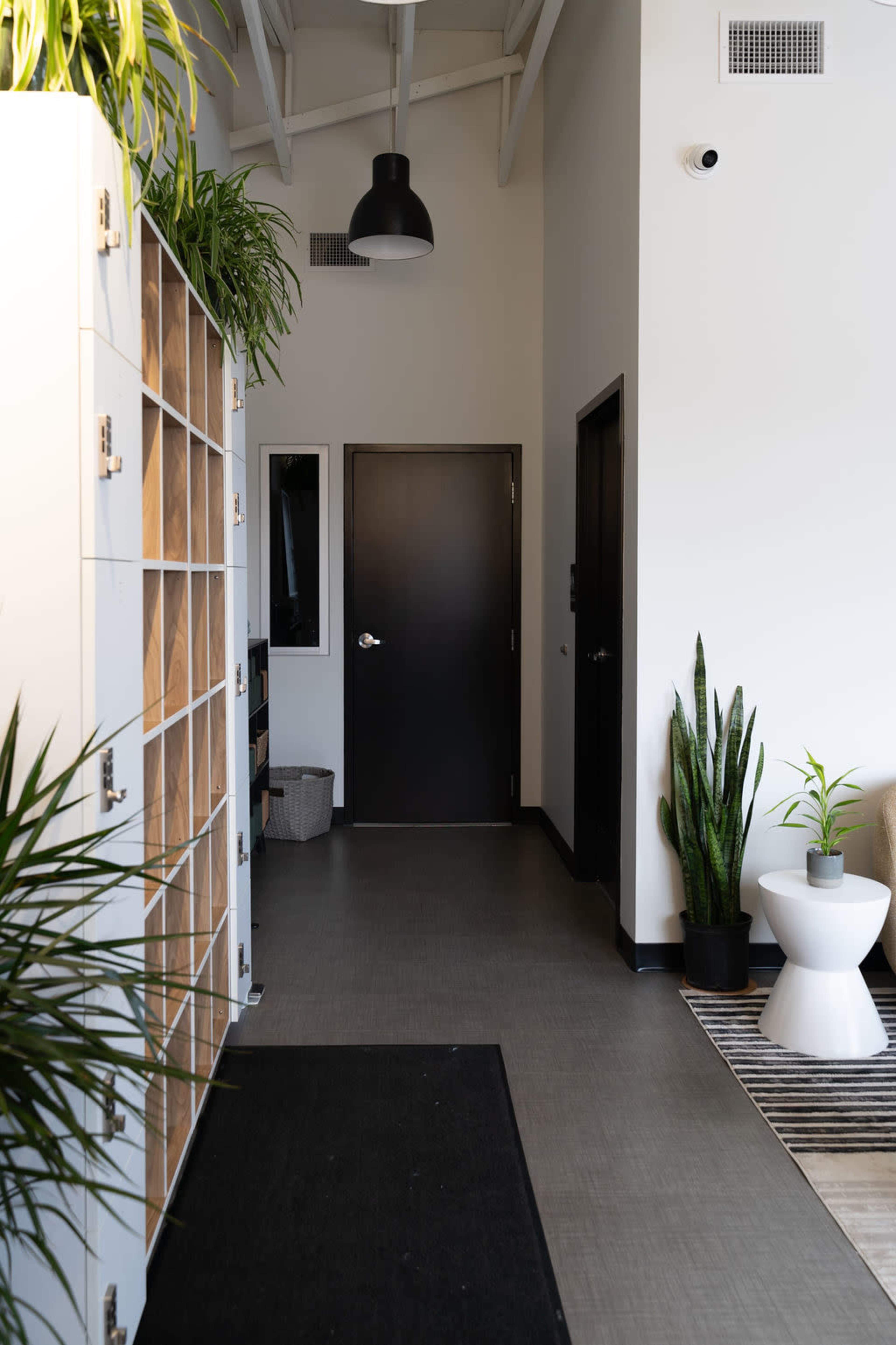 The image shows a modern hallway featuring a plant-filled entry area, lockers on one side, a black door at the end, and natural light illuminating the space.