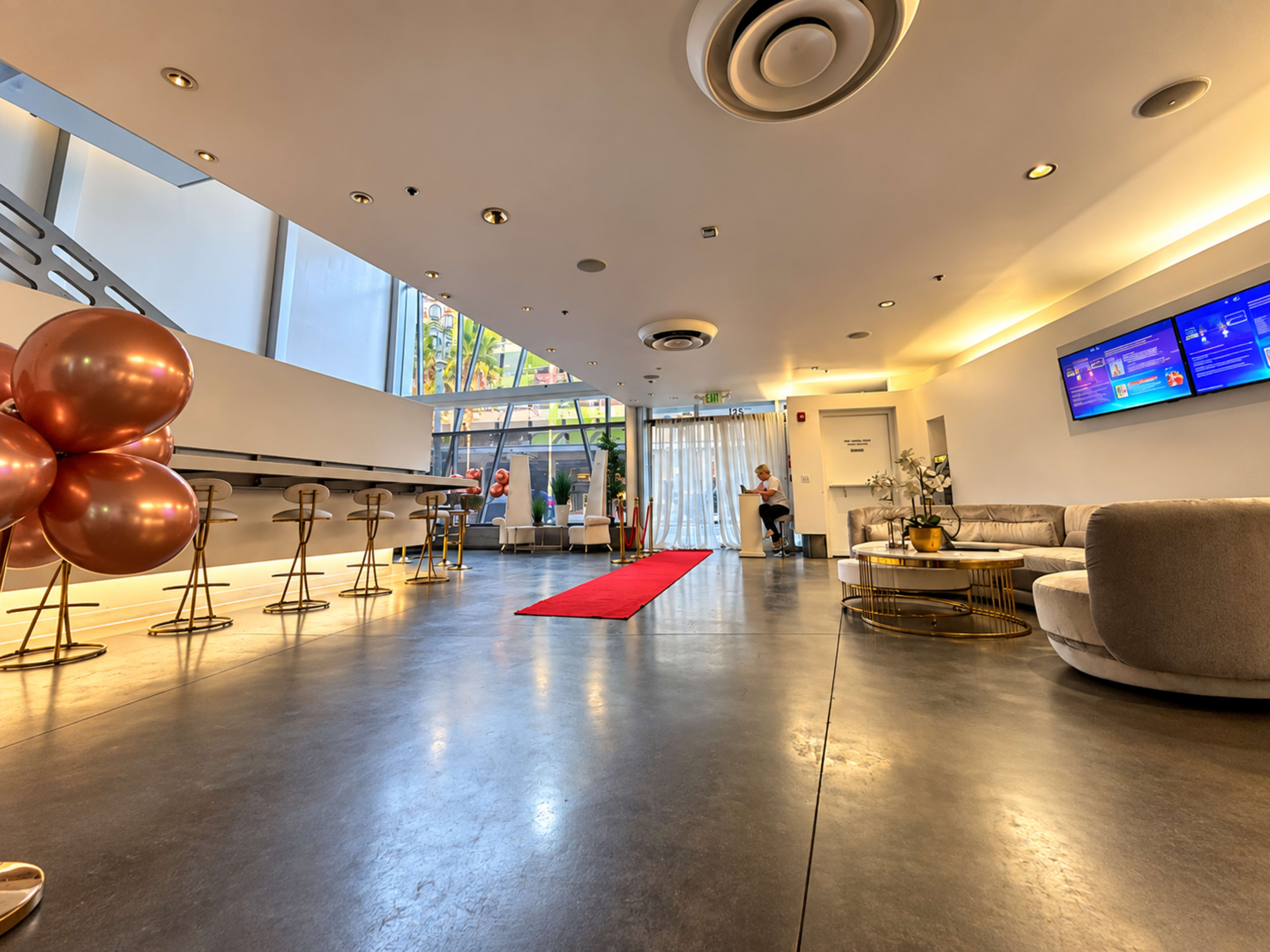 The image shows a modern interior lobby with a red carpet leading to a seating area and decorative golden balloons on one side.