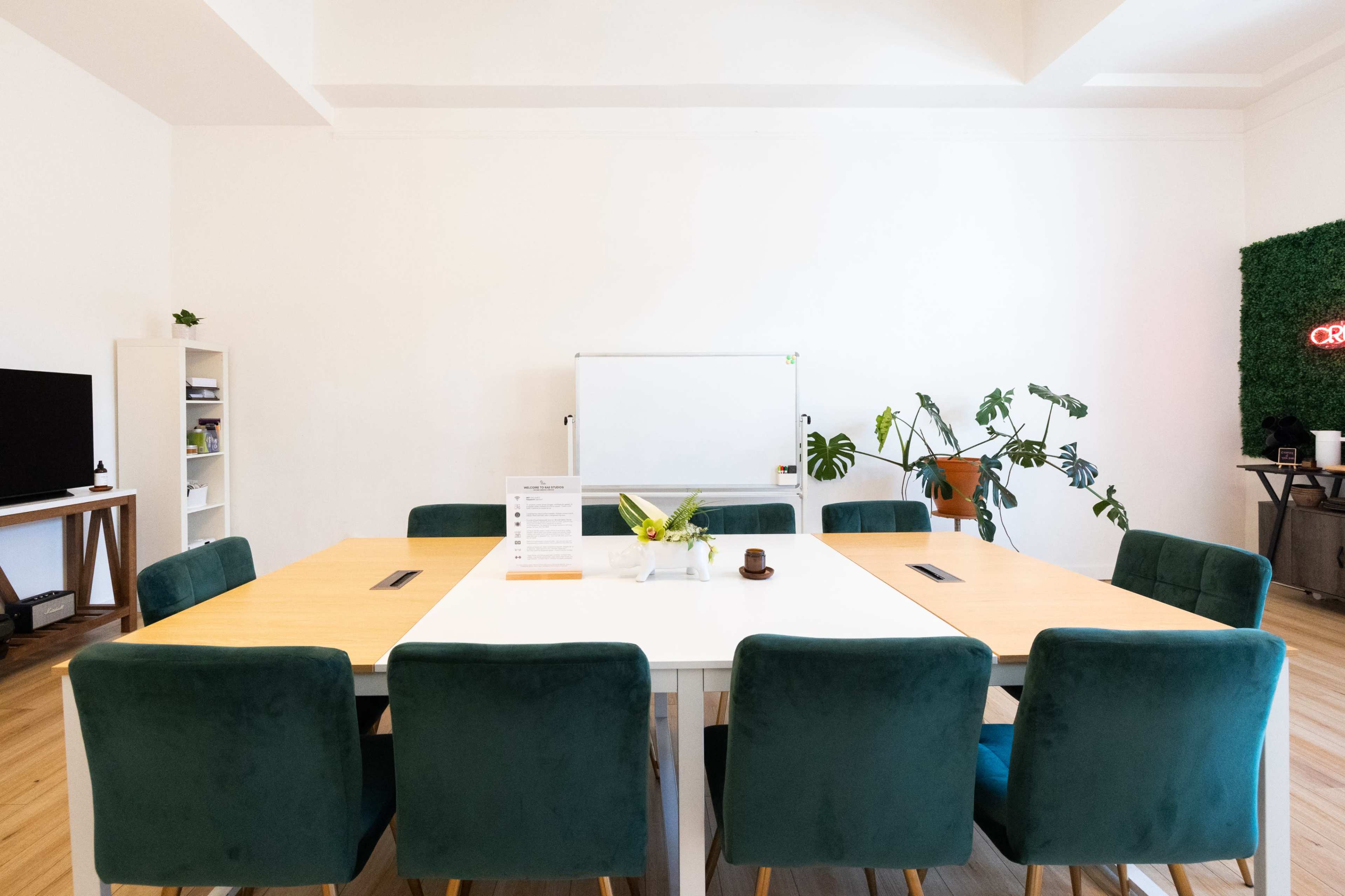 A conference room features a large table surrounded by green chairs, with a whiteboard and plants in the background.