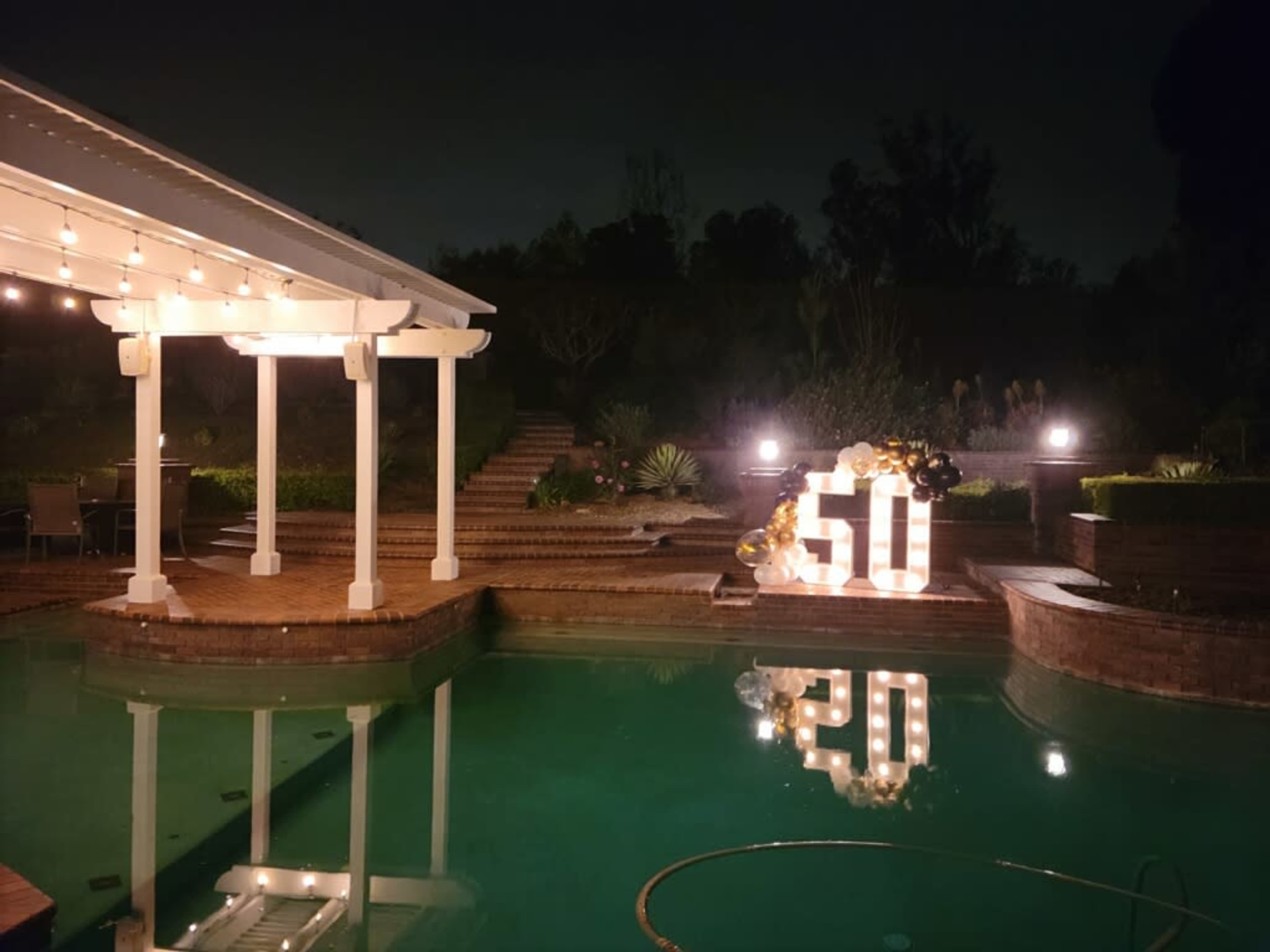 A brightly lit setup for a 50th birthday celebration is displayed near a pool at night, with decorative lights and flowers.