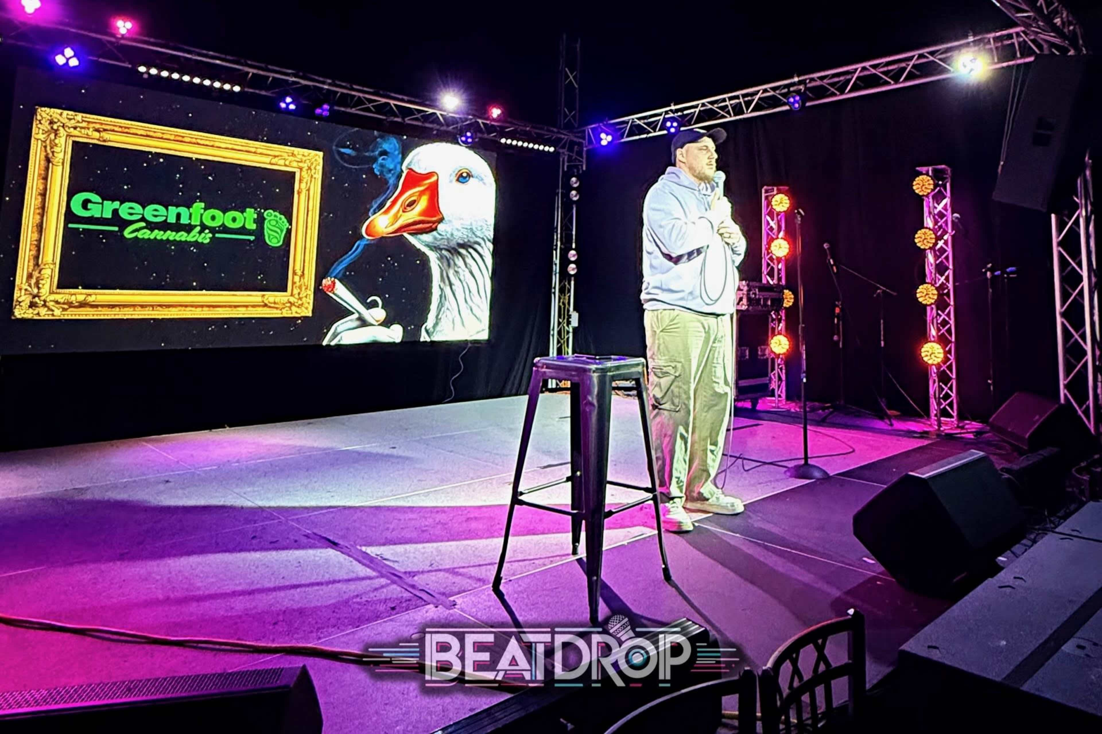 A comedian performs on stage in a venue featuring colorful lighting and a large screen displaying a graphic with the text "Greenfoot Cannabis" alongside a cartoon goose.
