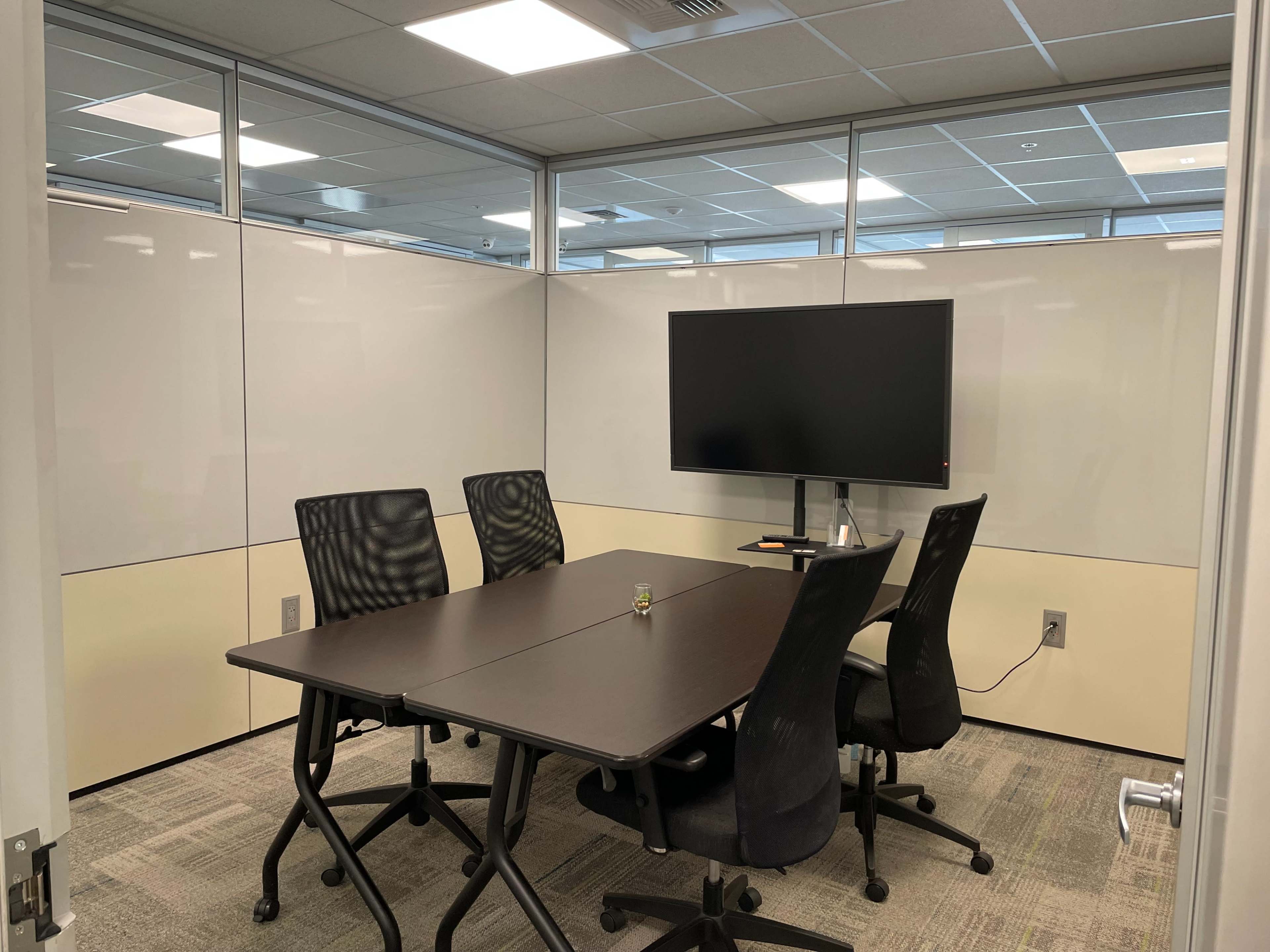 A small conference room with a table, four chairs, and a wall-mounted monitor.