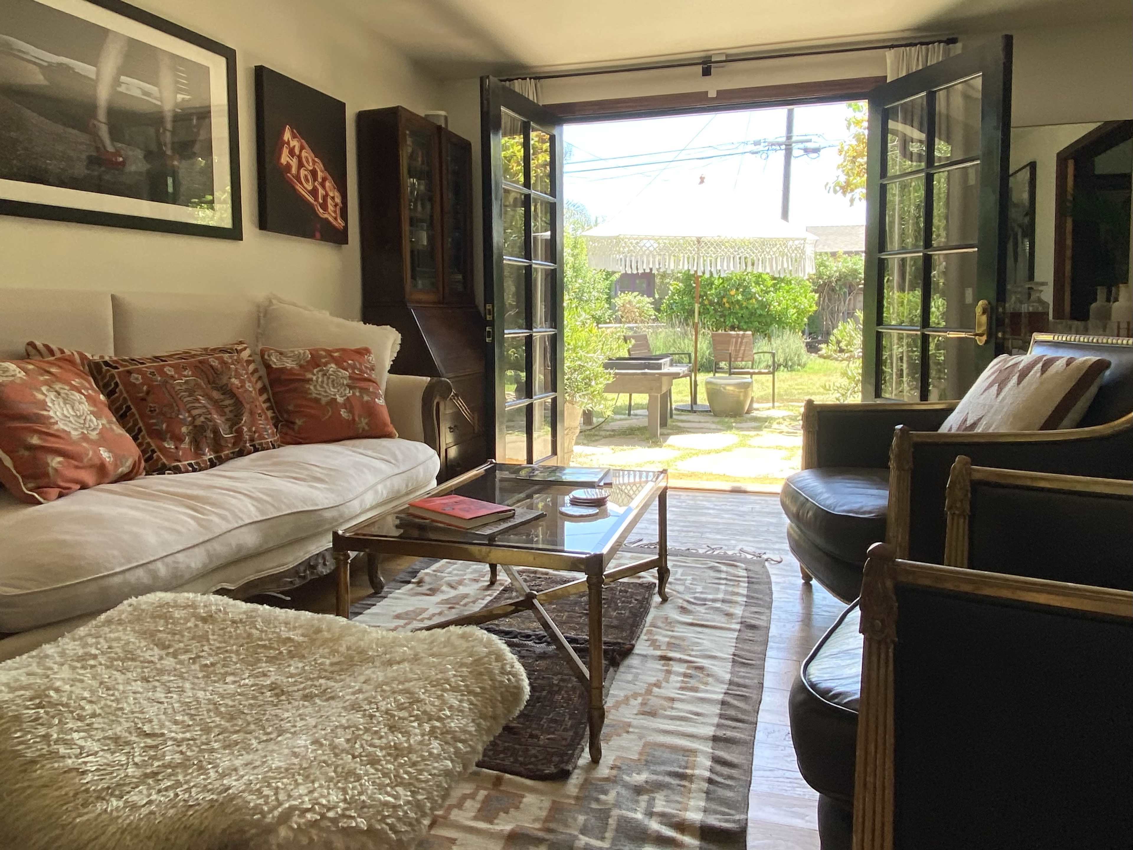The image shows a cozy living room with a patterned rug, a white sofa adorned with decorative pillows, and large open doors leading to a garden area outside.