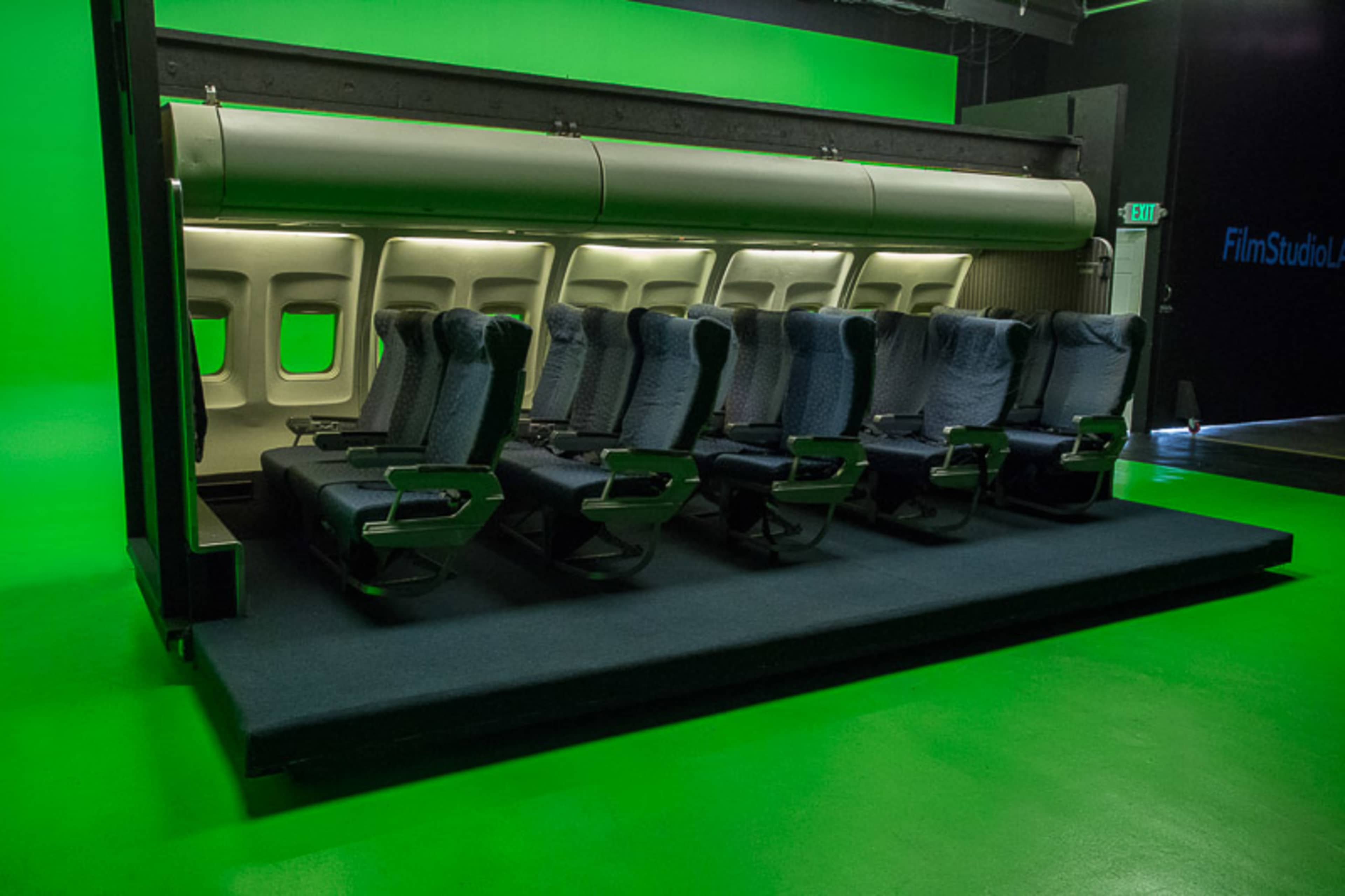 A row of airplane seats is positioned in front of a green backdrop in a film studio.
