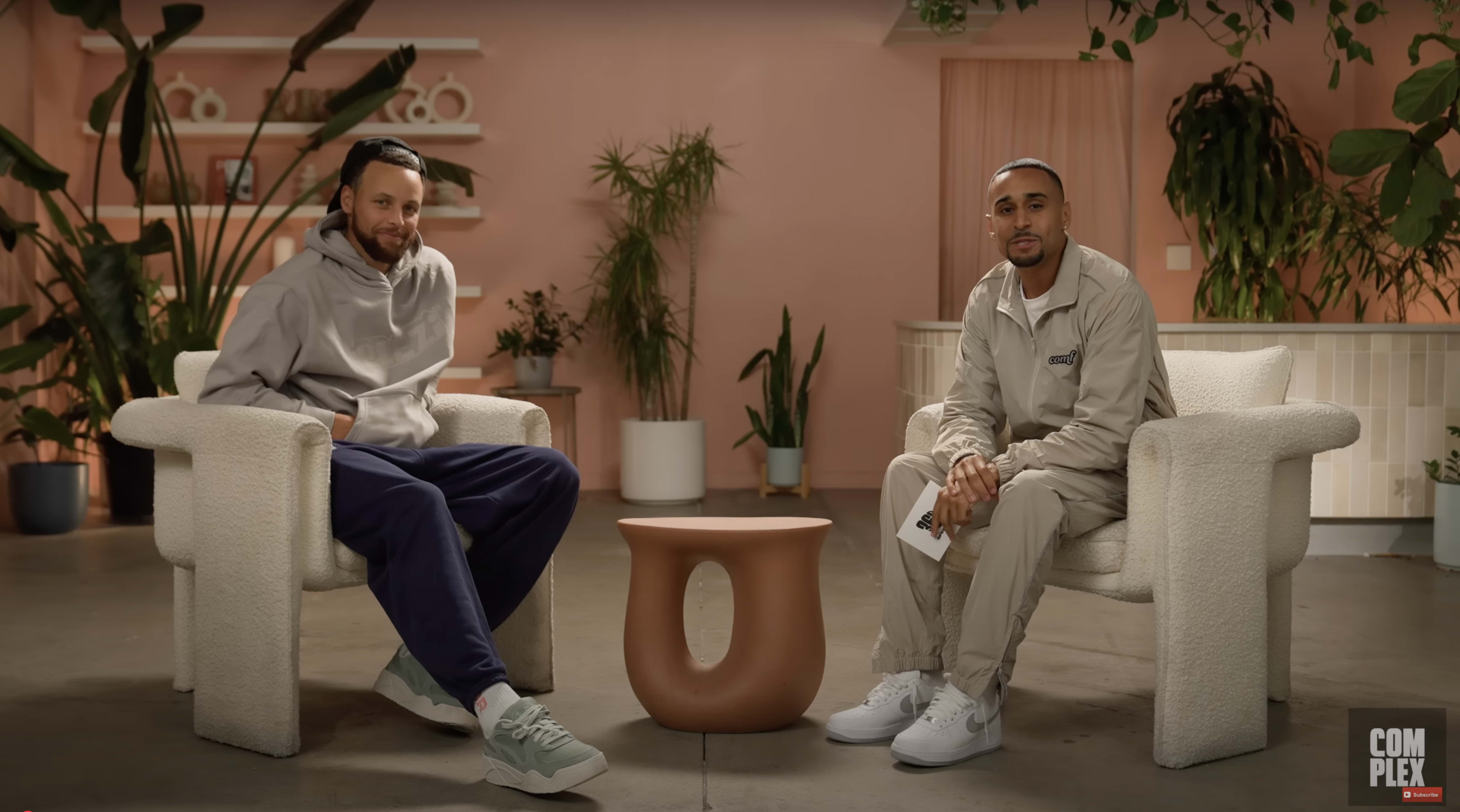 Two men sit in minimalist armchairs in a well-lit room with plants and a modern coffee table between them.