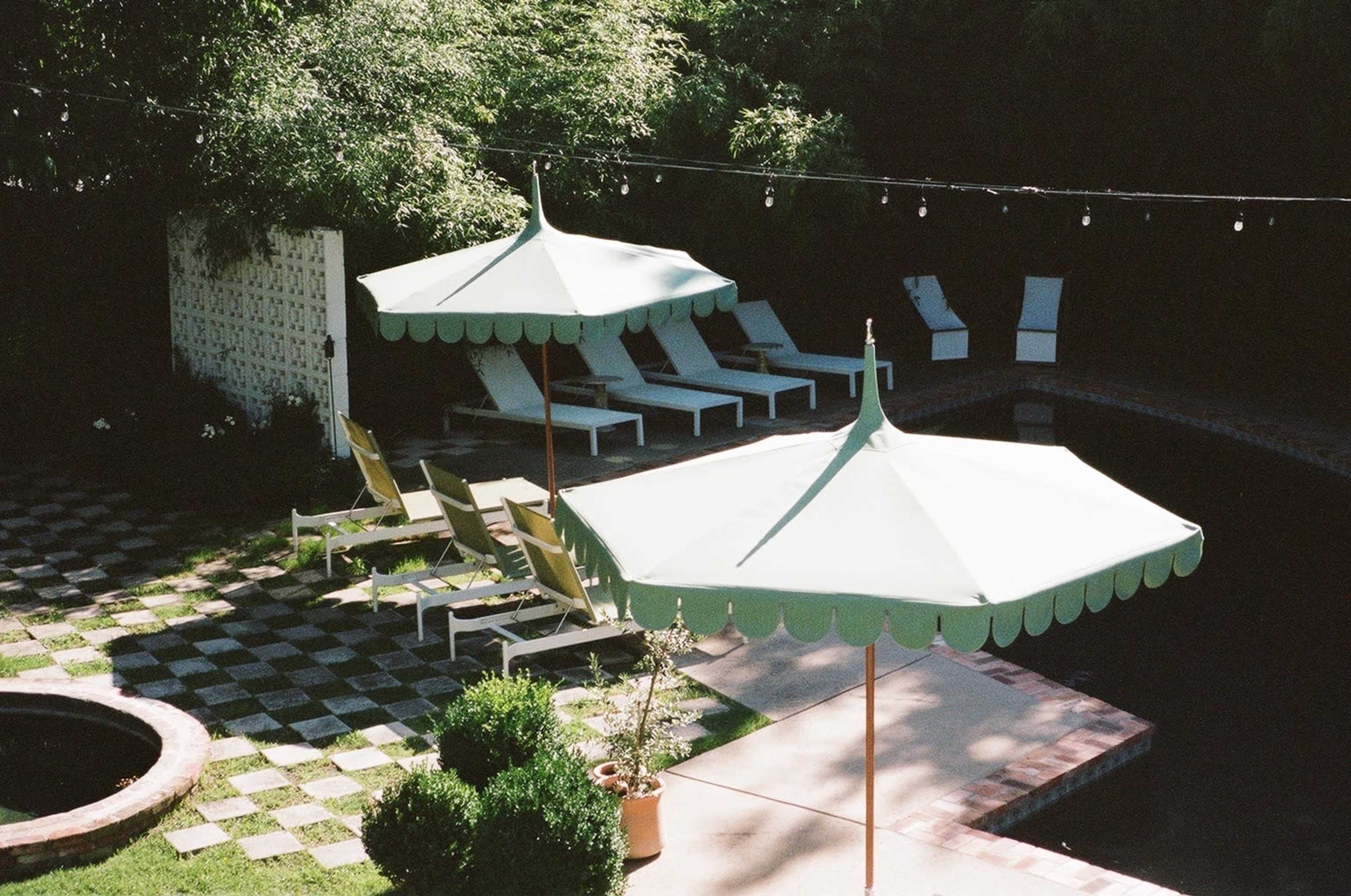 The image shows a pool area with two green umbrellas shading lounge chairs, surrounded by a checkered stone pattern and potted plants.