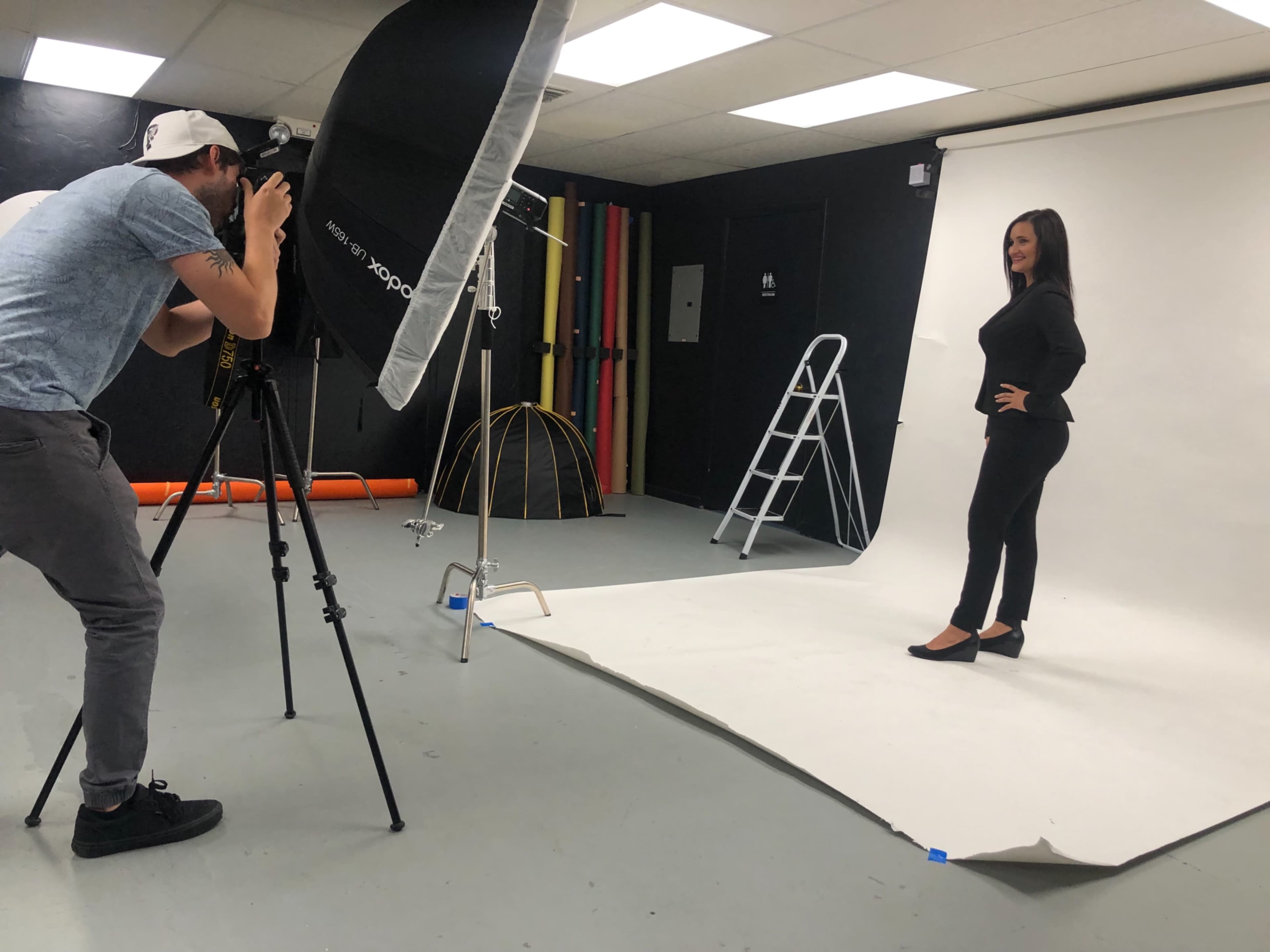A photographer is capturing a portrait of a woman posing in a studio against a white backdrop.