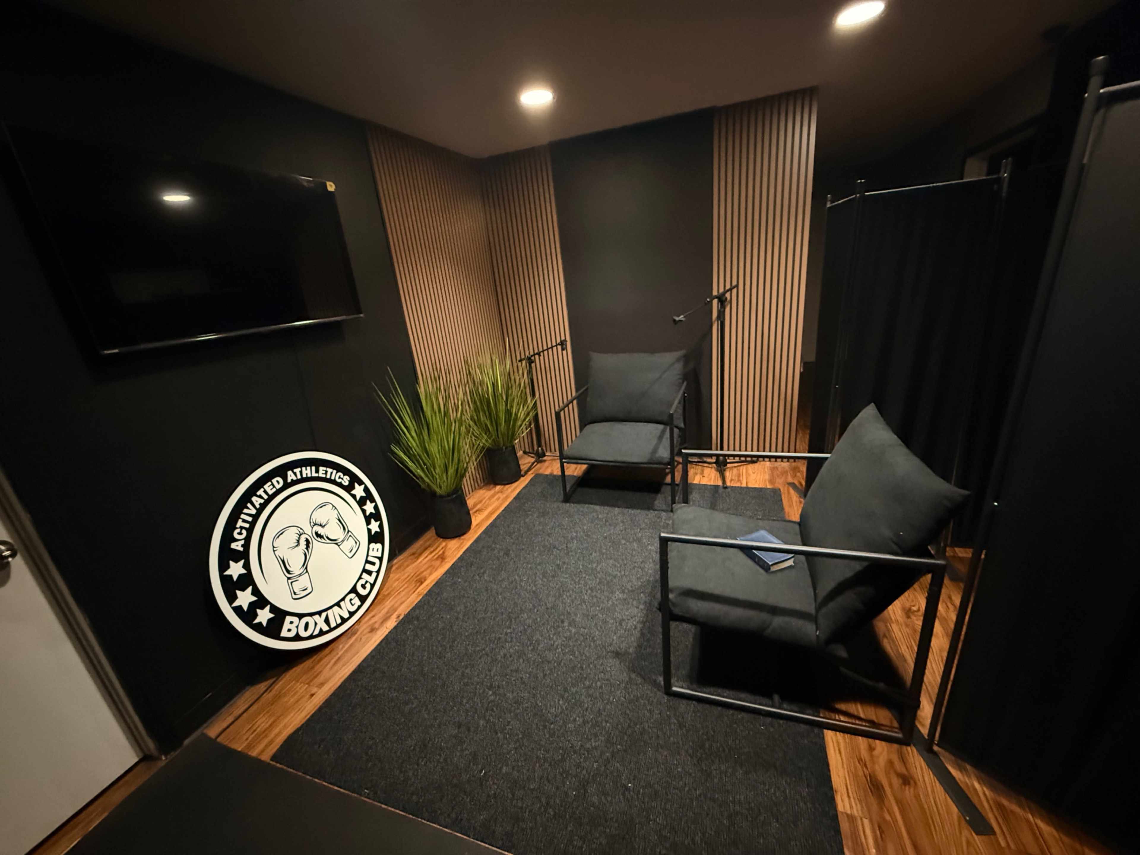 The image shows a small, modern room with two black chairs, a textured wall, a television mounted on the wall, and a logo for a boxing club on the floor.