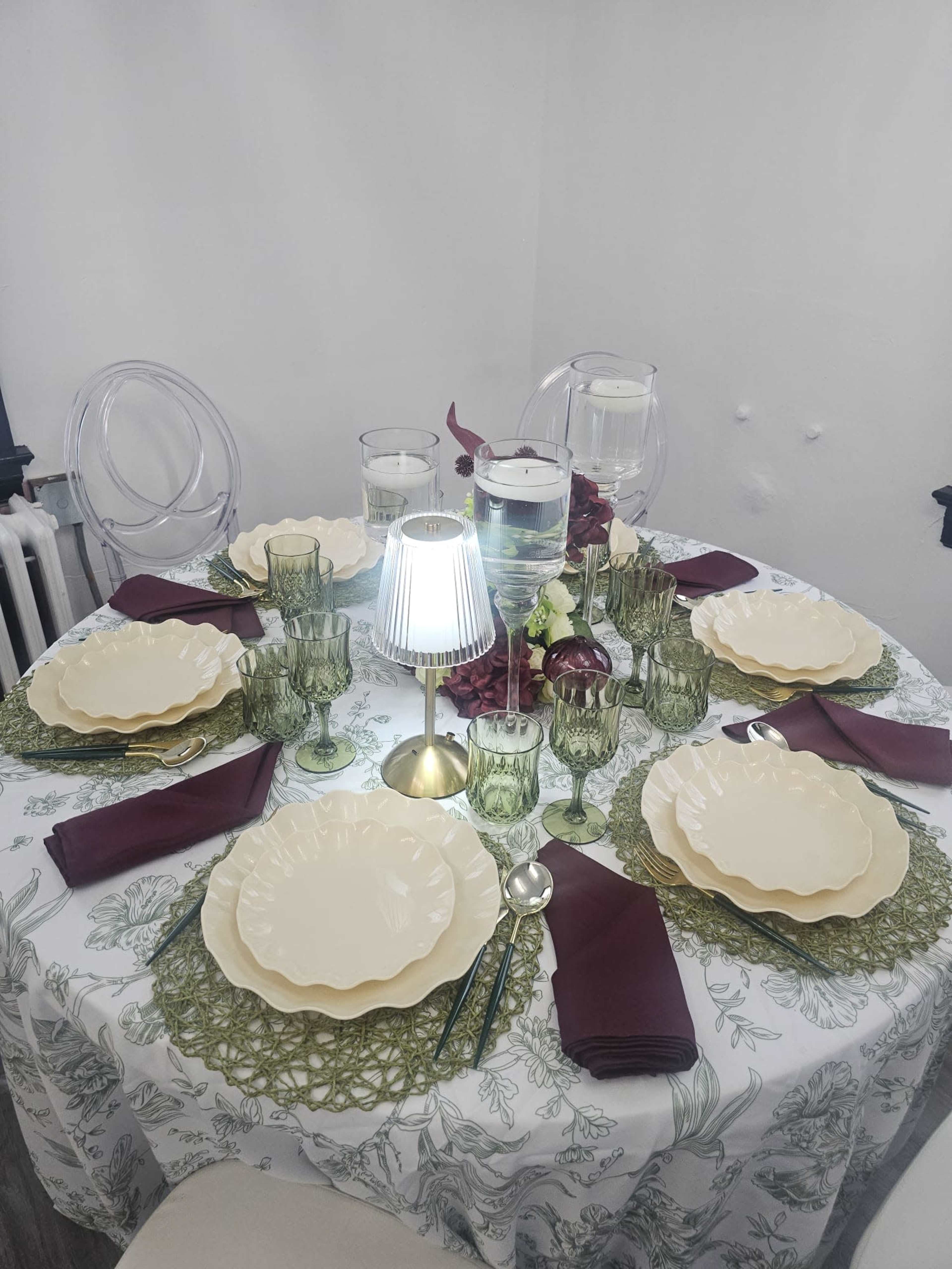 A round dining table is elegantly set with green and white tableware, featuring a floral tablecloth, layered placemats, and illuminated by a small lamp.