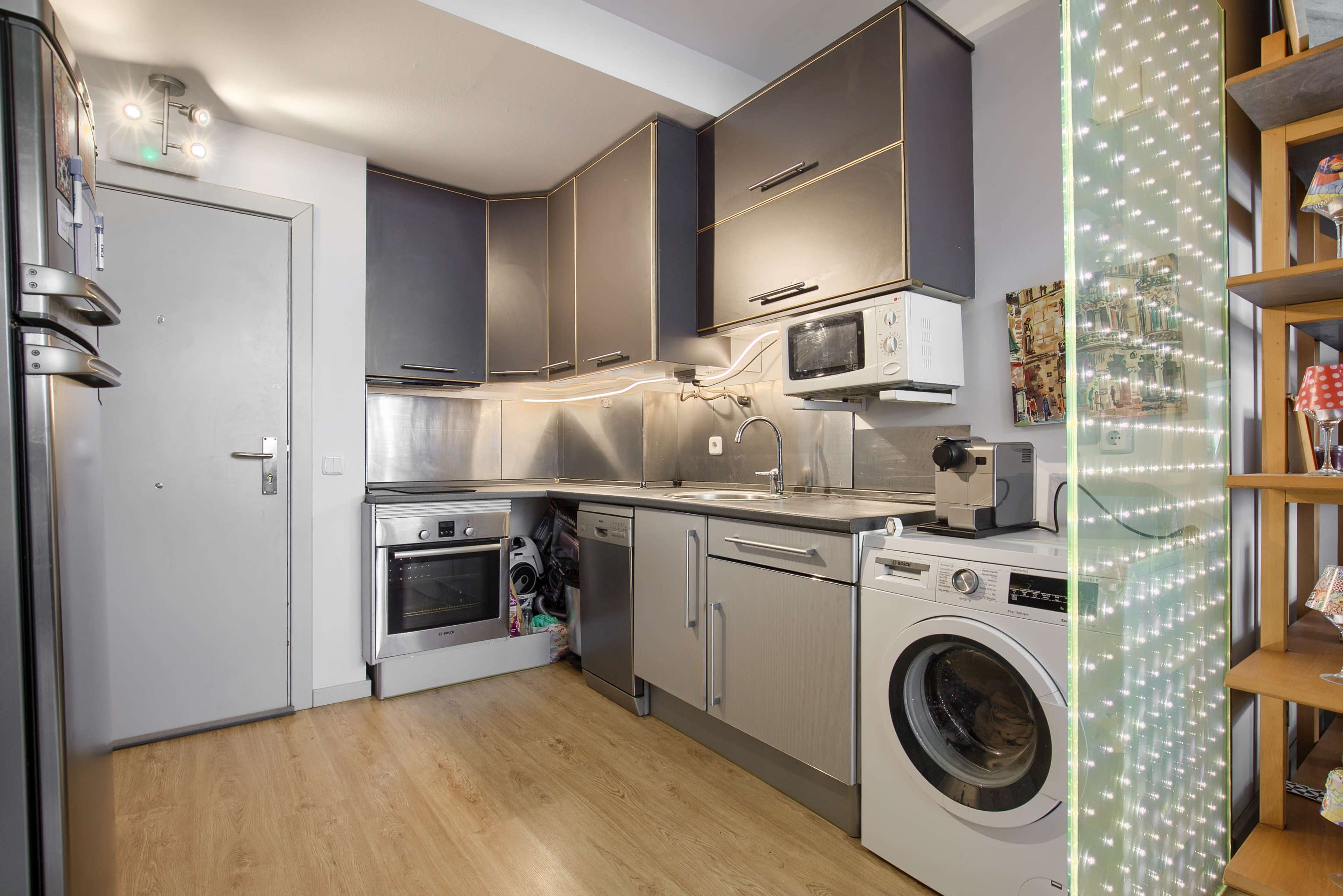 A compact kitchen featuring modern appliances, including a refrigerator, oven, microwave, and washing machine, with a lighted decorative panel on the right.