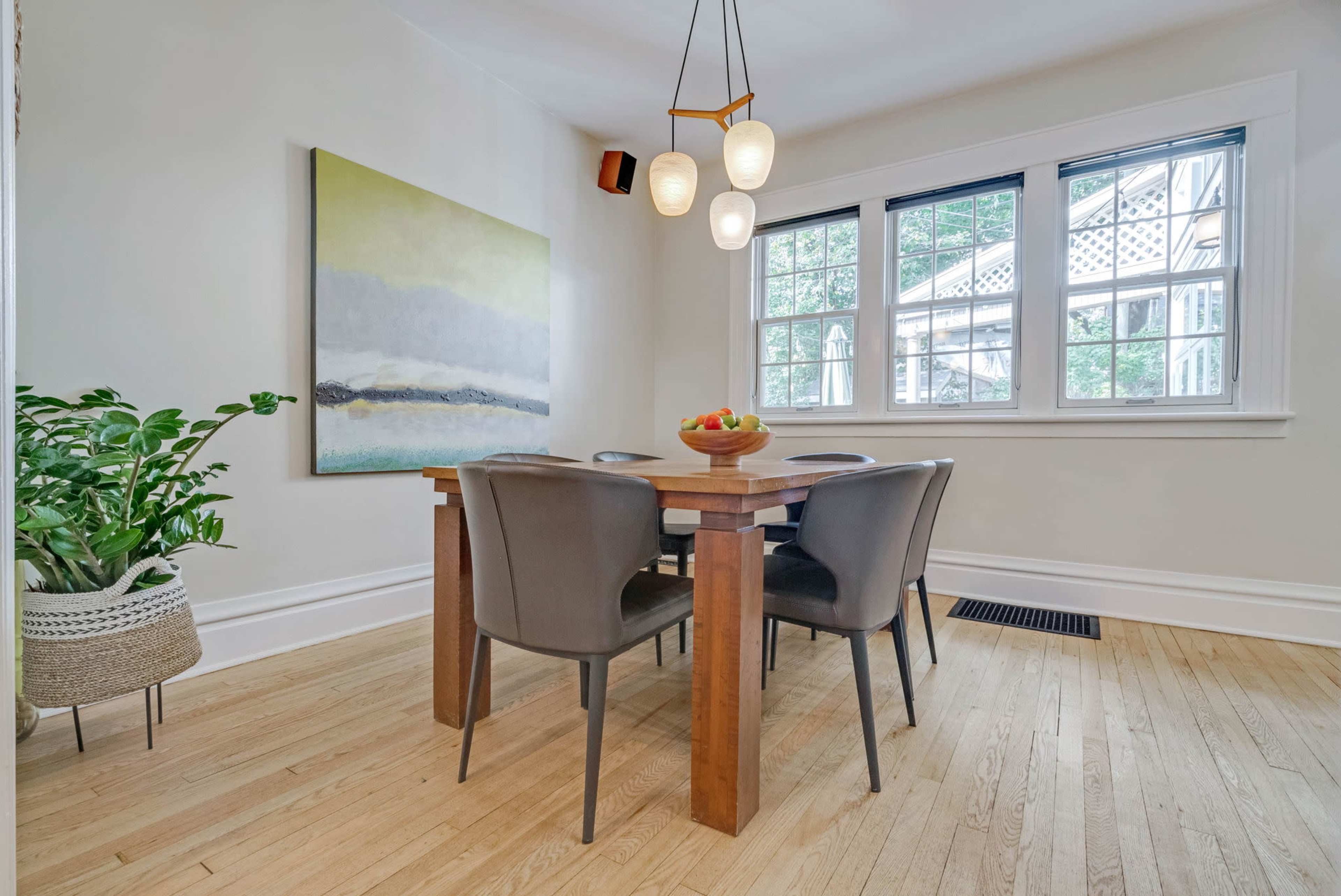 A modern dining area features a wooden table surrounded by four chairs, a colorful painting on the wall, and a plant in a basket.