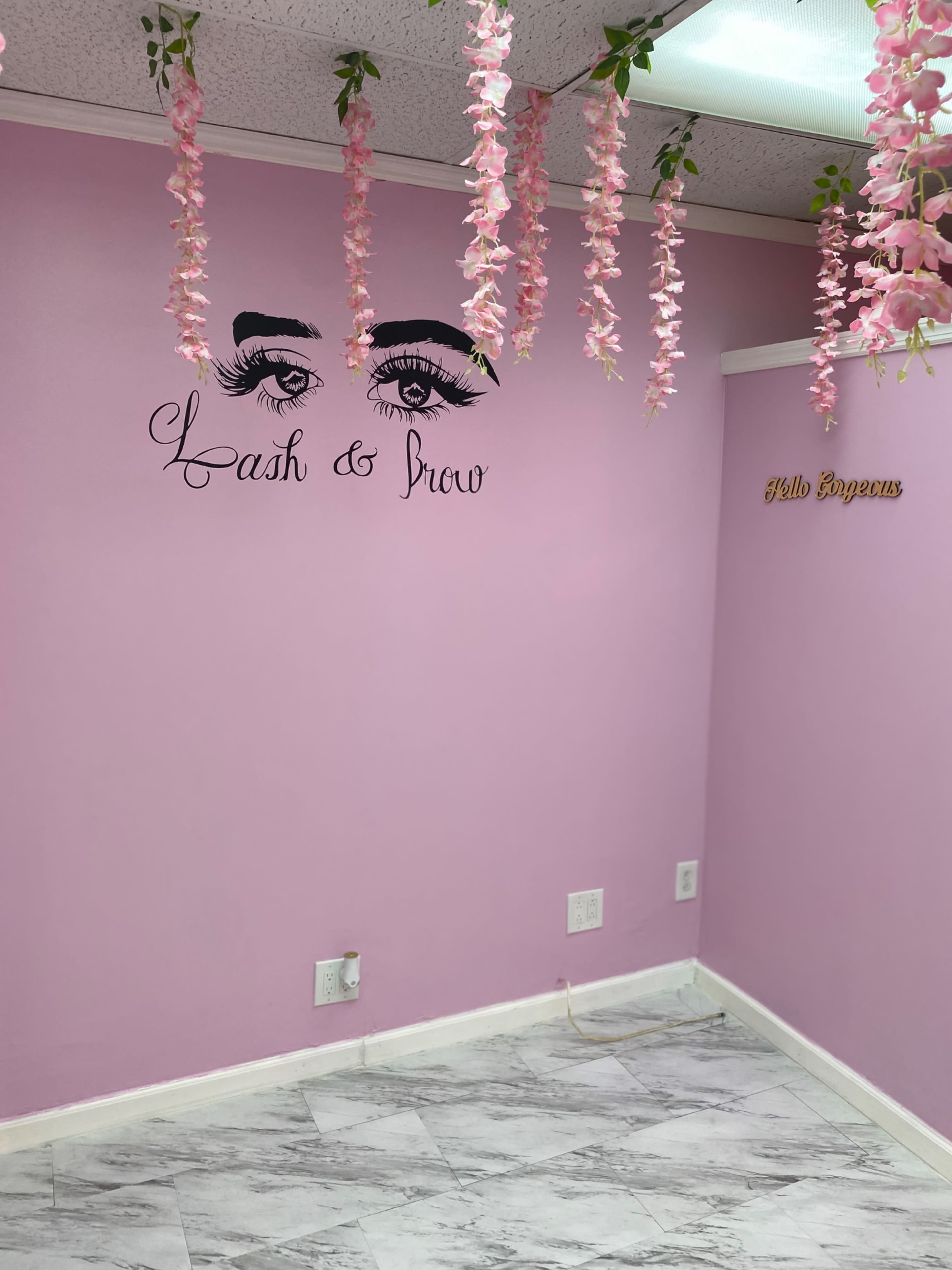 A pink-walled room with a decorative wall featuring the phrase "Lash & Brow" and artificial flowers hanging from the ceiling.