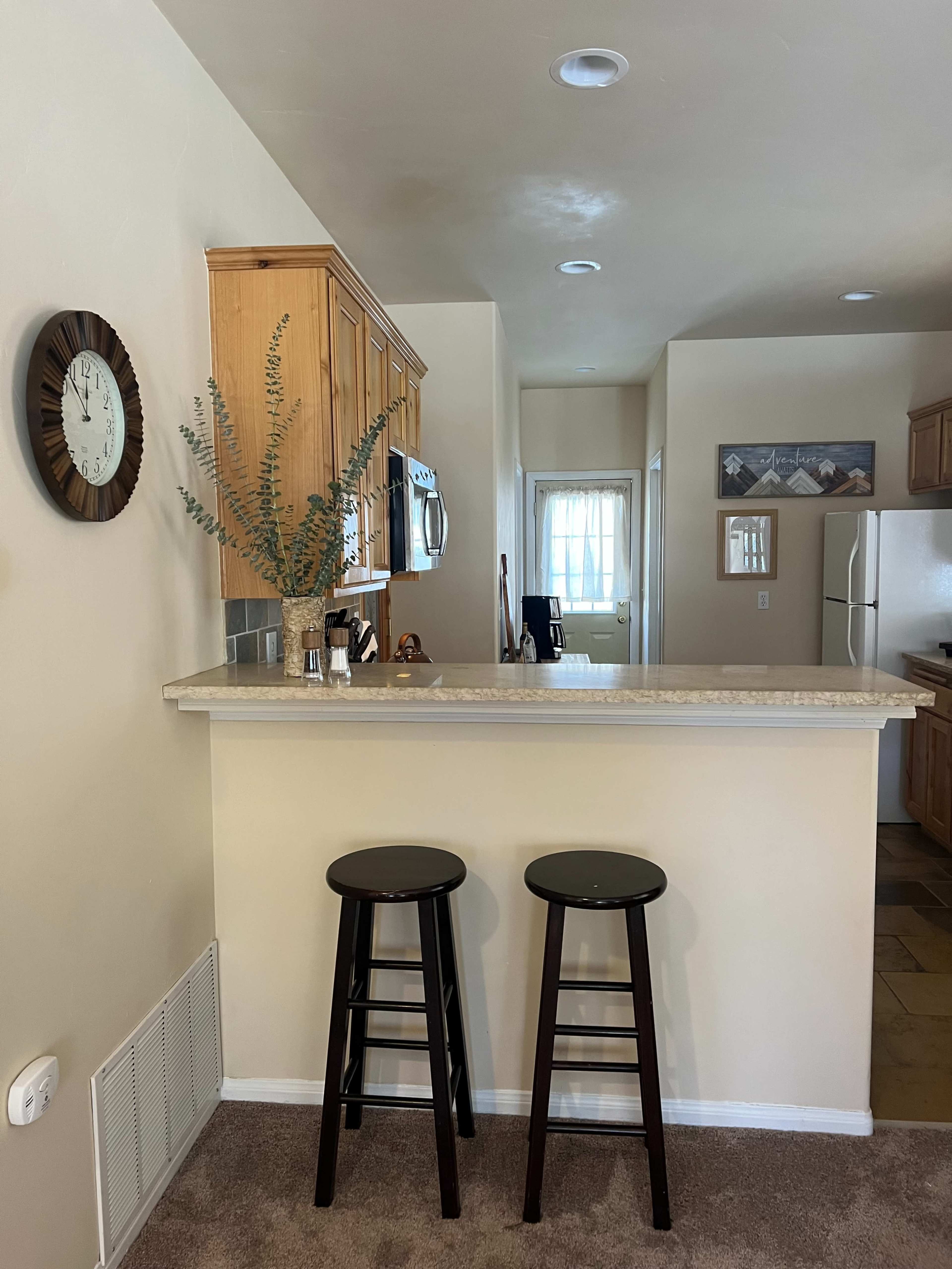 A kitchen area with a counter, two black stools, wooden cabinets, and a clock on the wall.