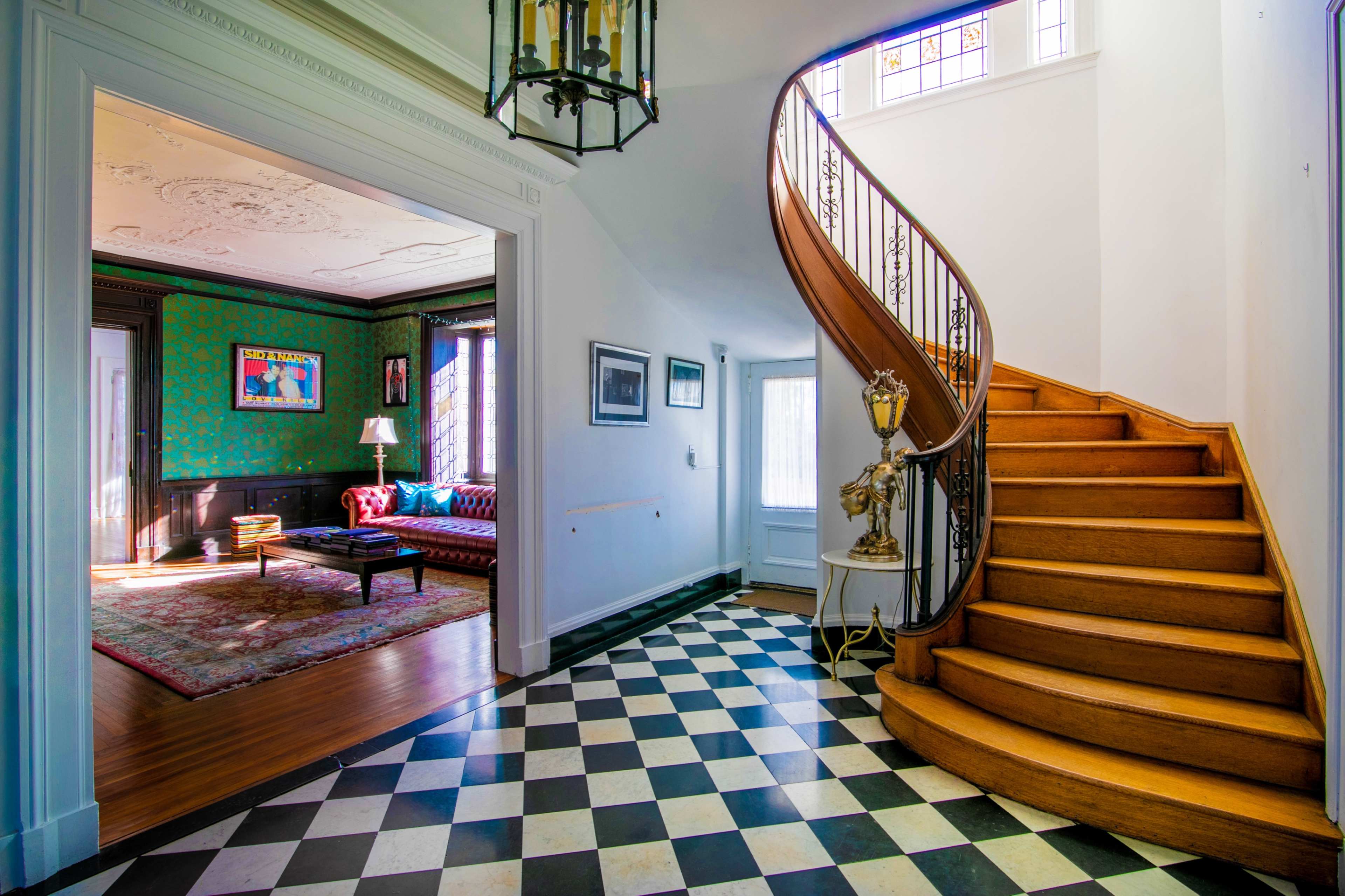The image features an entryway with a black and white tiled floor leading to a wooden staircase, with a glimpse of a richly decorated living room visible through an open door.