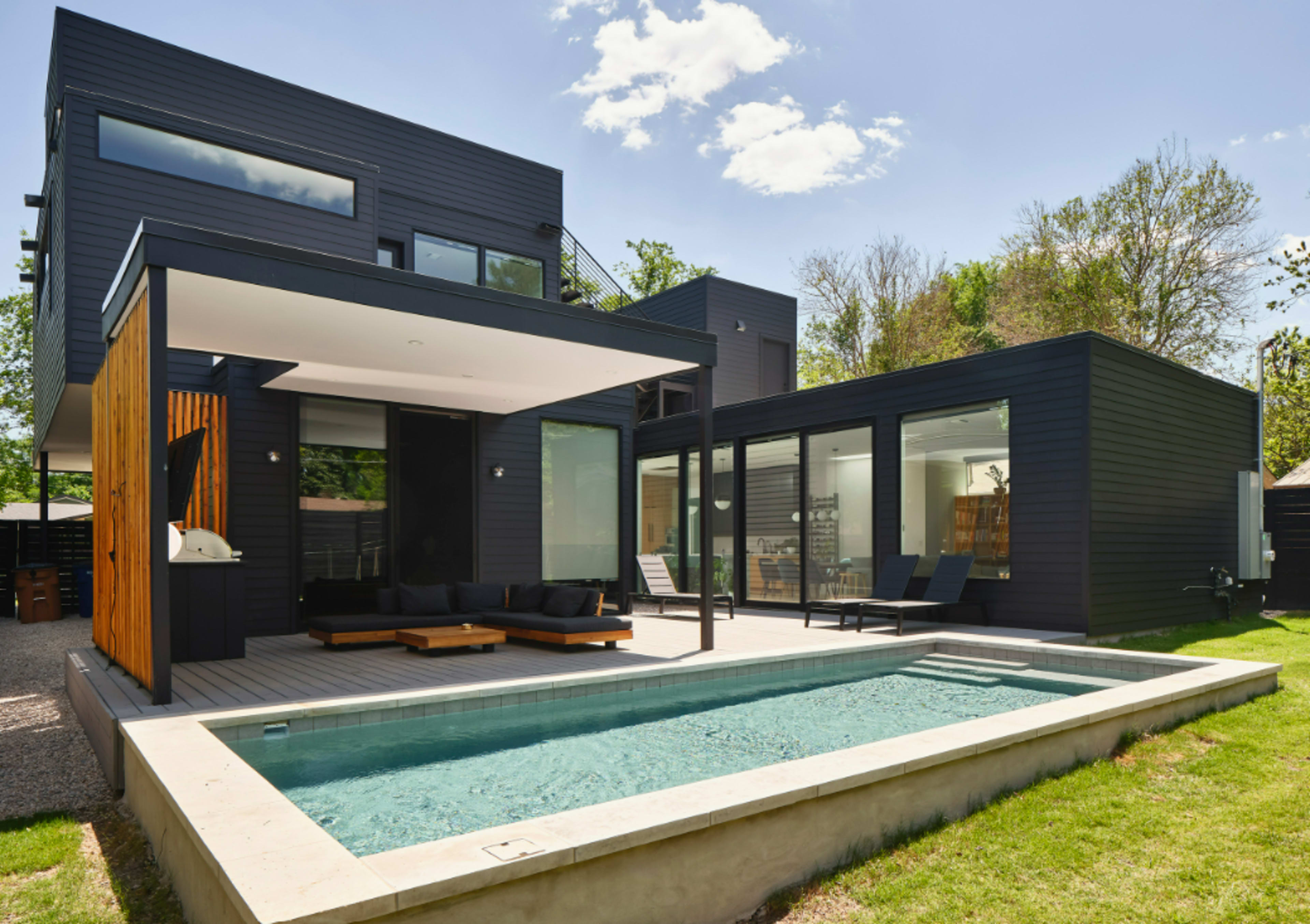 The image shows a modern house with a rectangular swimming pool in the foreground and large windows that allow ample natural light.