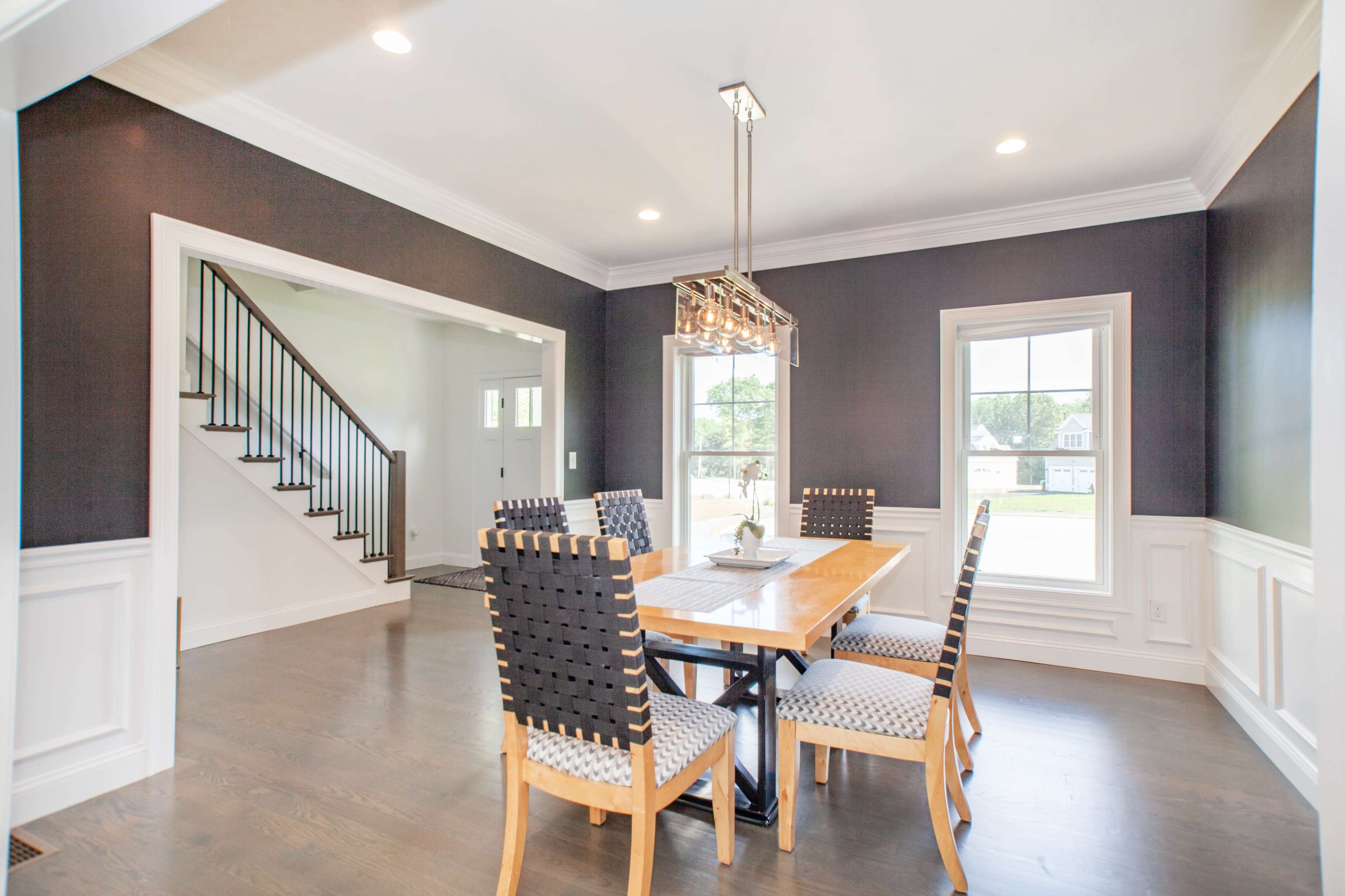 A dining area features a wooden table surrounded by six chairs, with a staircase visible in the background and large windows providing natural light.