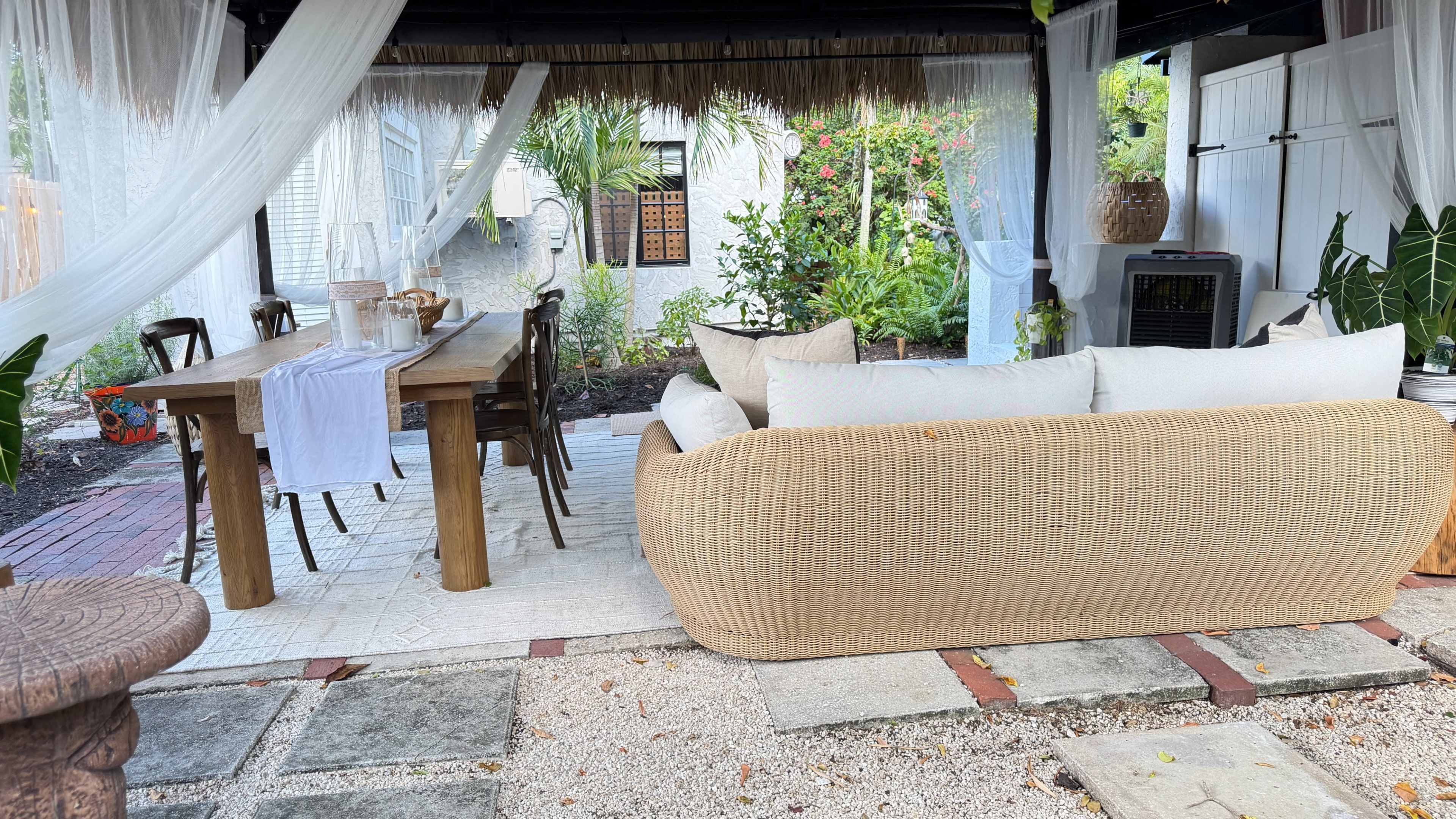 The image shows a covered outdoor seating area with a large table, a couch, and surrounding greenery.