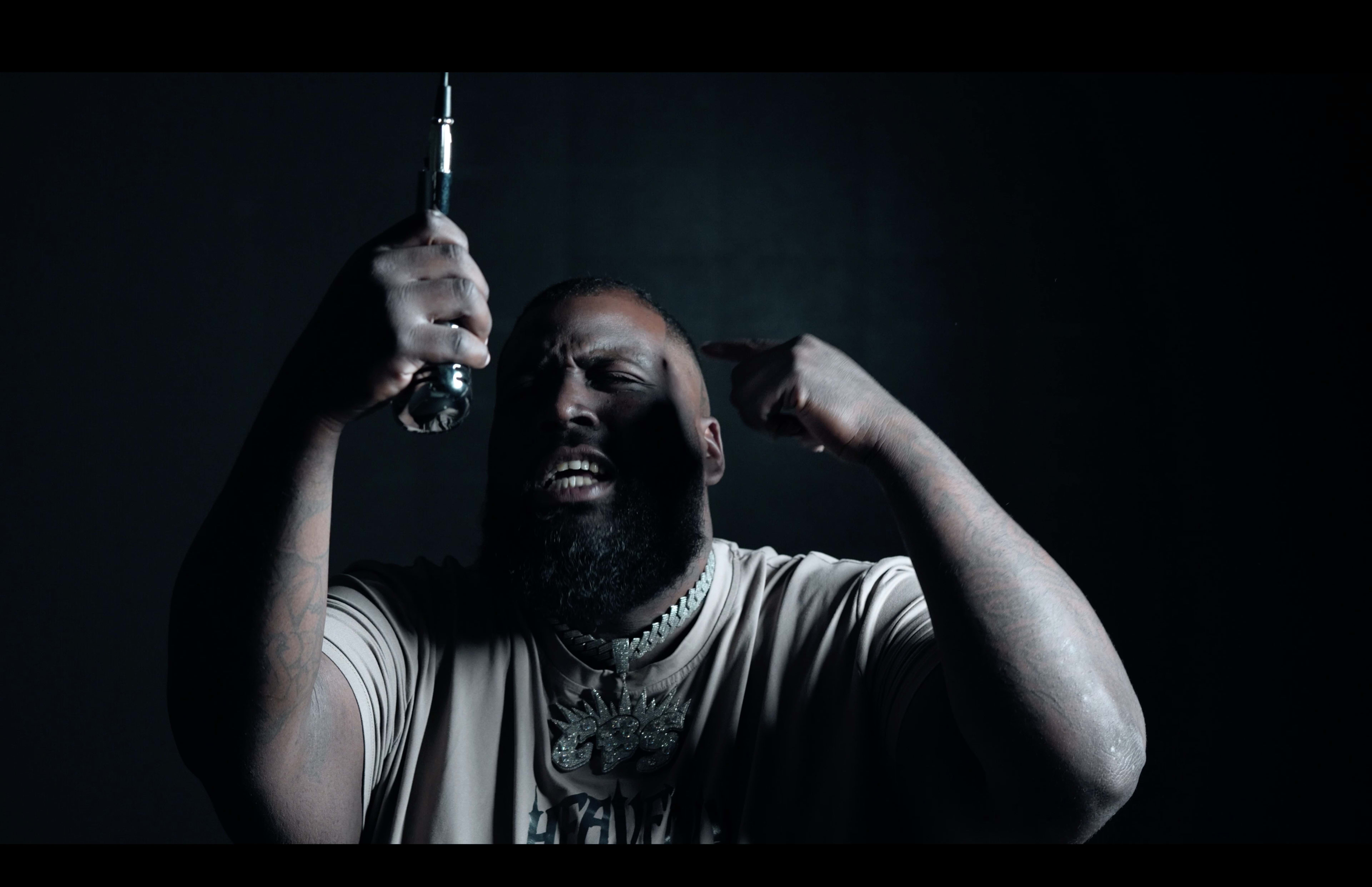 A man with a beard and tattoos passionately holds a microphone while gesturing with his other hand against a dark background.