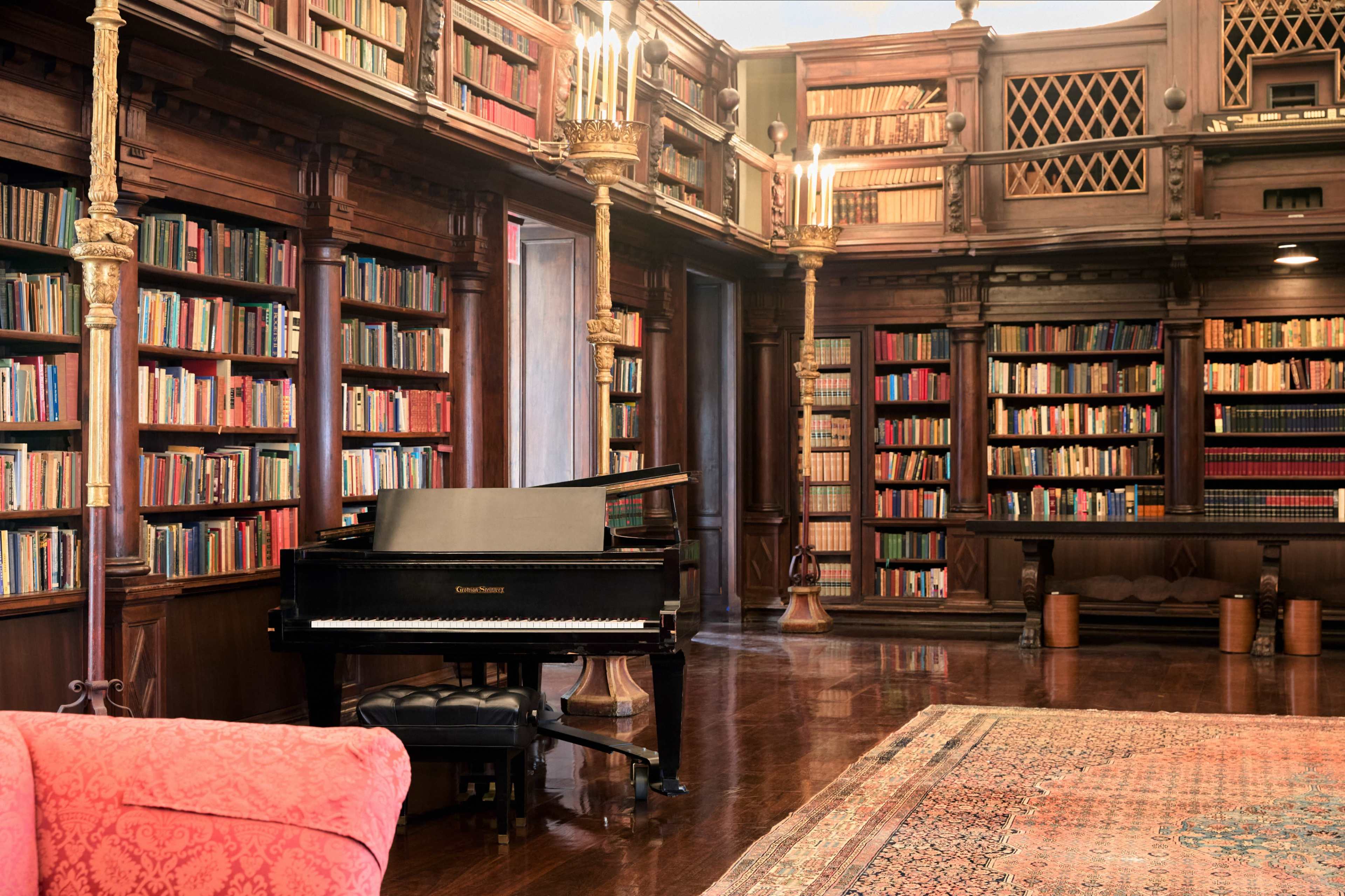 The image shows a well-appointed library featuring ornate wooden bookshelves filled with books, a grand piano positioned in the corner, and a richly patterned area rug on the floor.