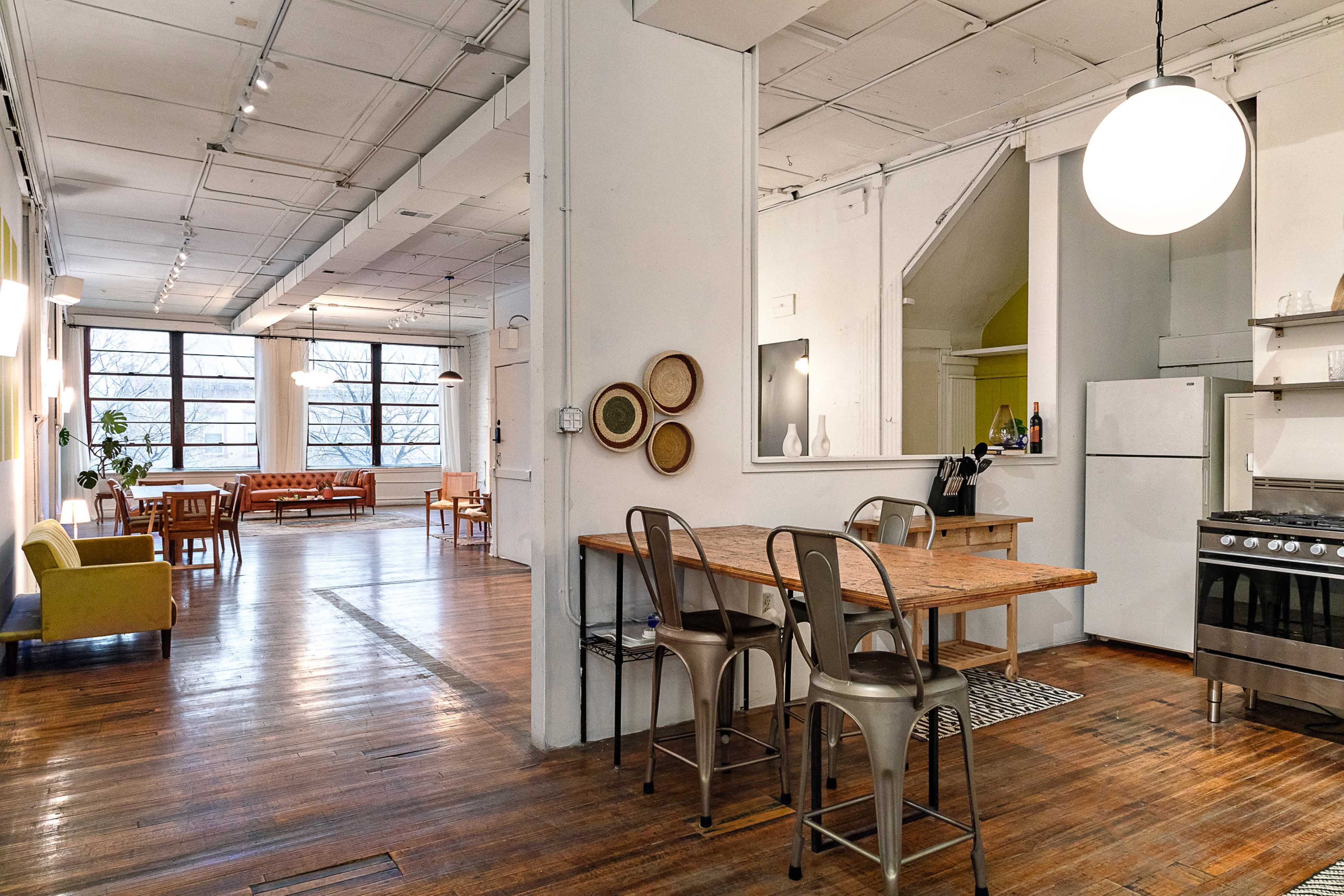 An open-concept living space features a kitchen area with a wooden dining table and metal stools, leading to a bright lounge area with large windows and minimalist furniture.