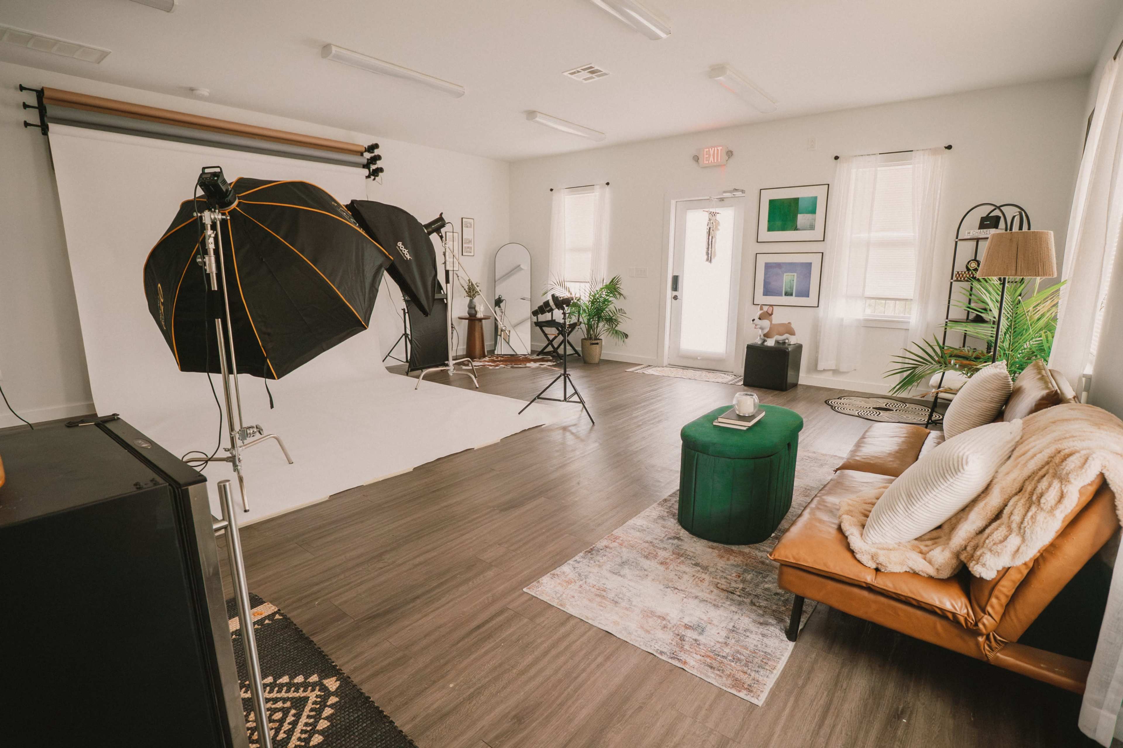 The image shows a well-lit photography studio featuring a leather couch, a green ottoman, and professional lighting equipment against a backdrop of a white wall.
