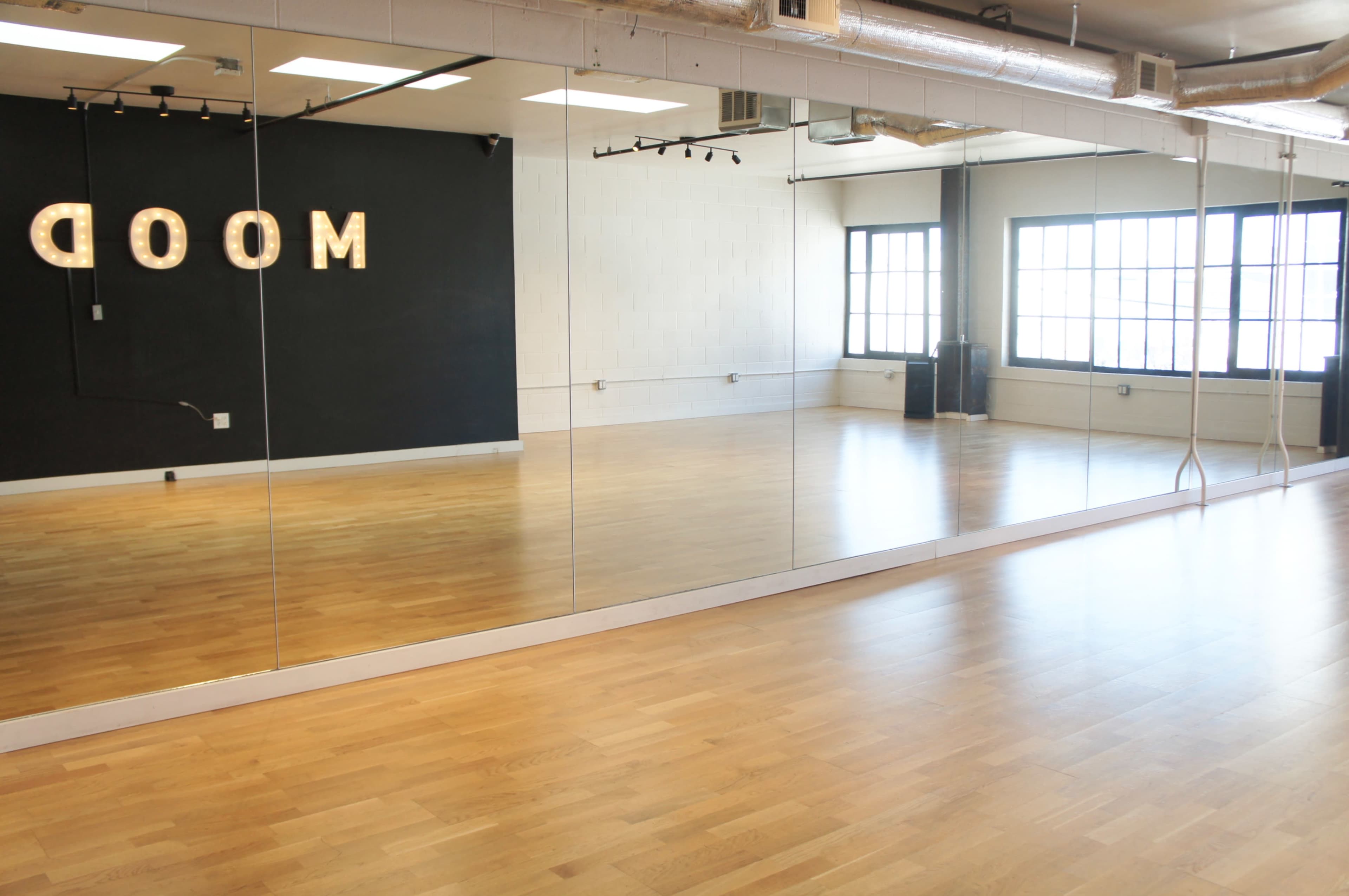 The image shows a spacious dance studio with polished wooden floors, large mirrors along one wall, and a dark backdrop featuring illuminated letters spelling "MOOD."