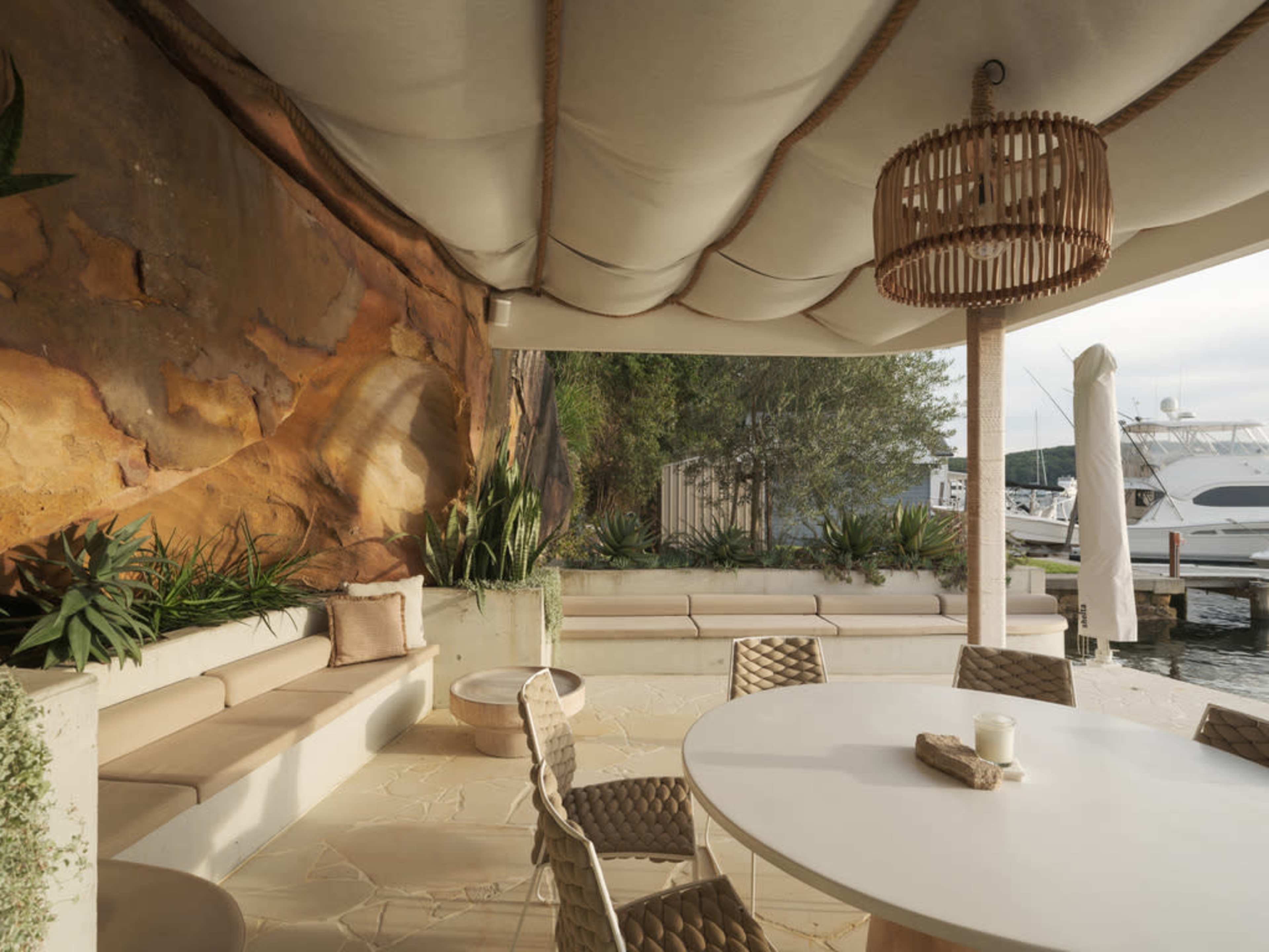 A covered outdoor seating area features a round table and cushioned benches against a natural rock backdrop, with boats visible in the background.