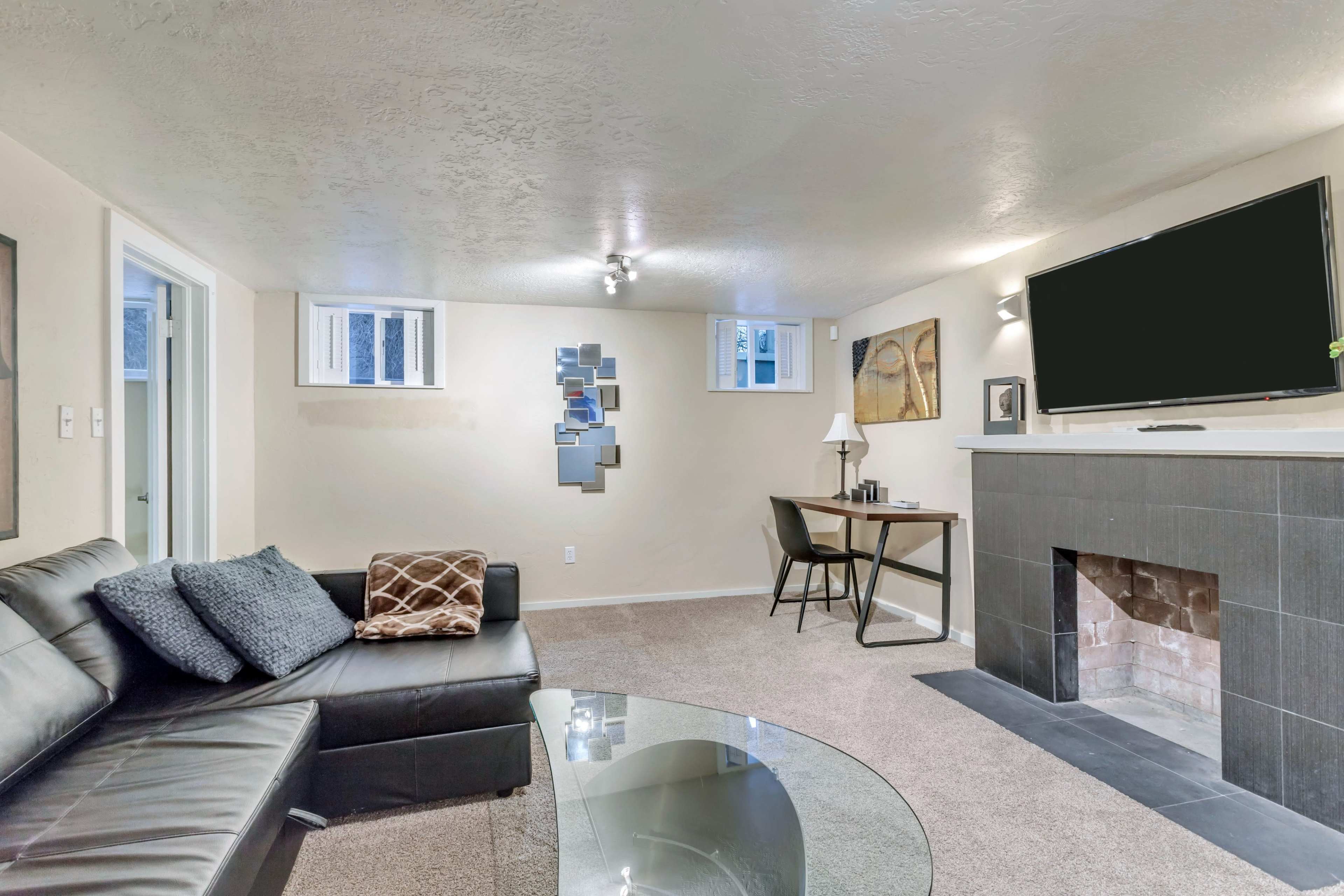 A well-lit basement living area featuring a black leather sectional sofa, a glass coffee table, a work desk with a lamp, and a flat-screen TV mounted above a stone fireplace.