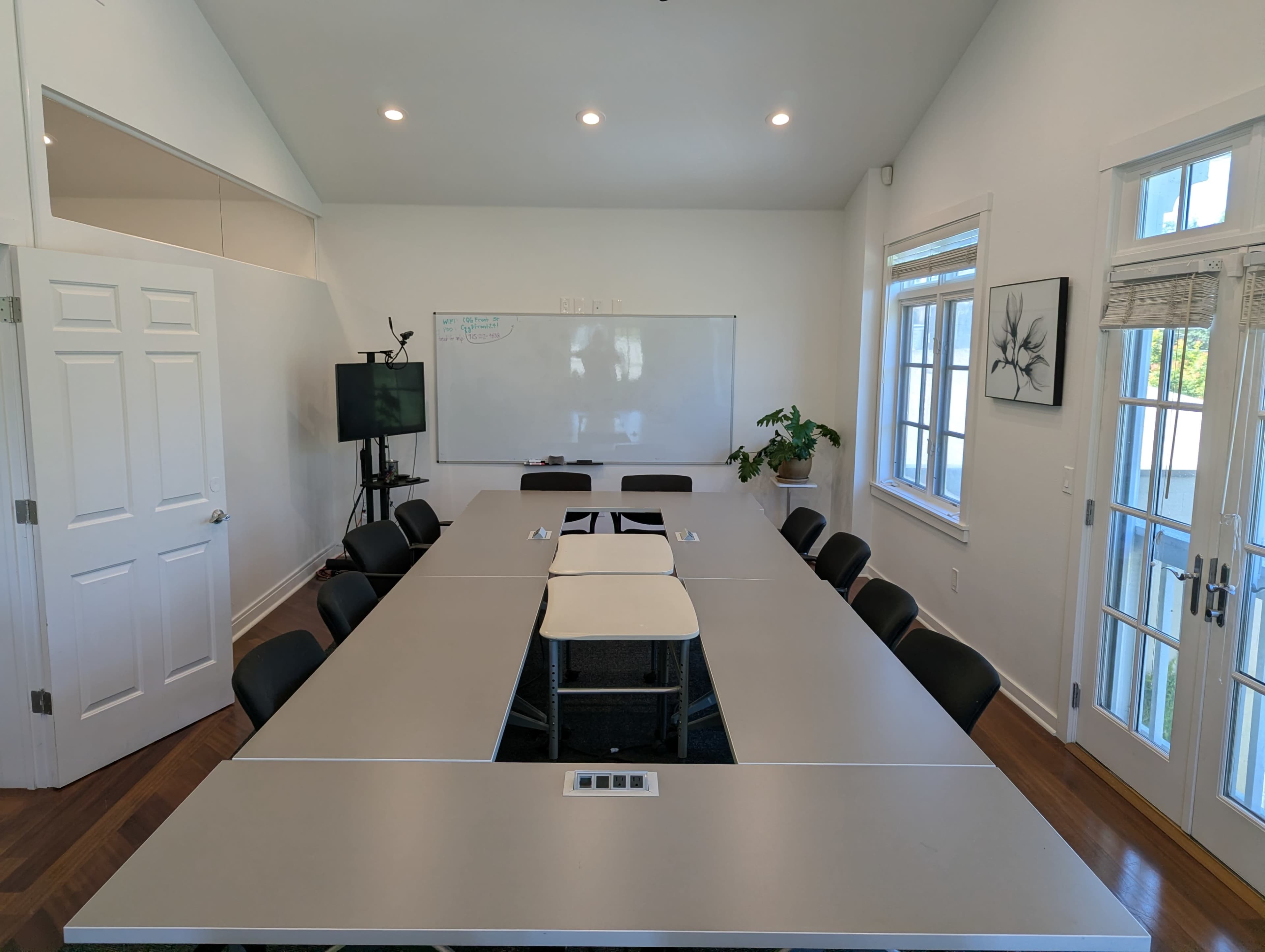 A conference room features a long table surrounded by black chairs, with a whiteboard on one wall and a plant in the corner.