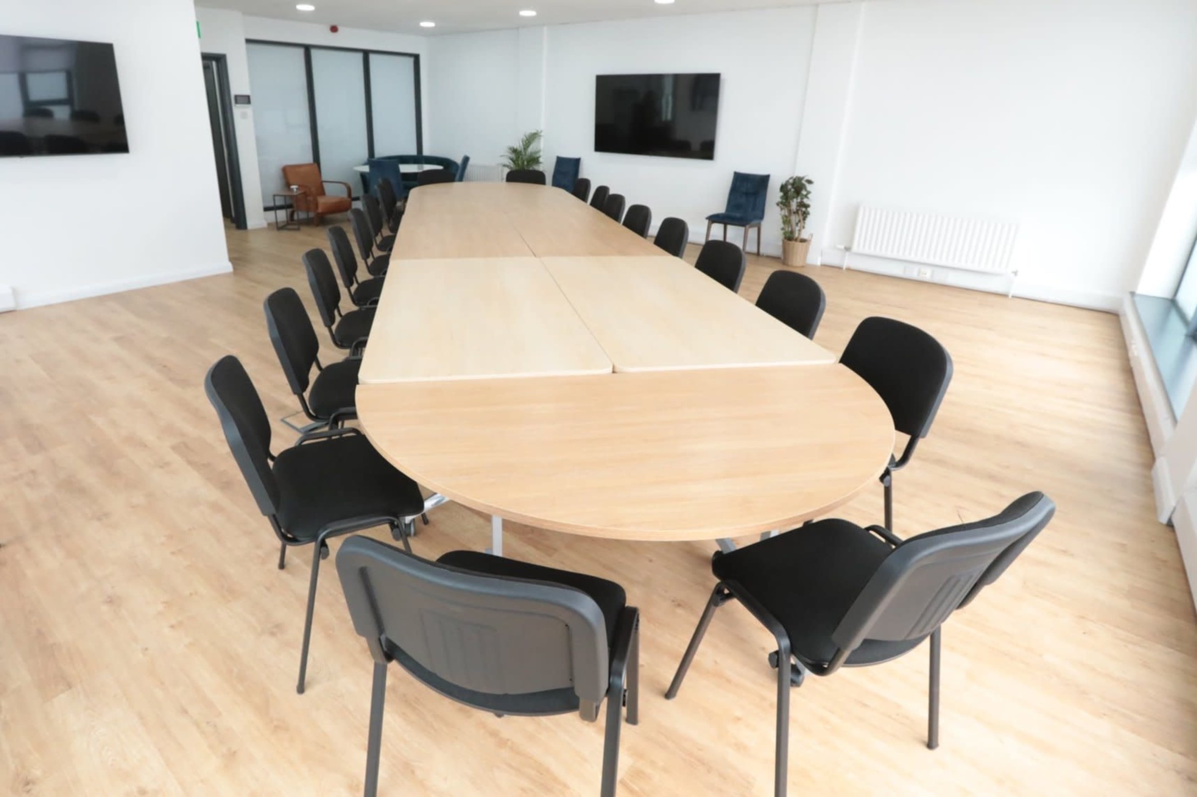 A large, oval conference table surrounded by black chairs is arranged in a bright, modern meeting room with wood flooring and large windows.