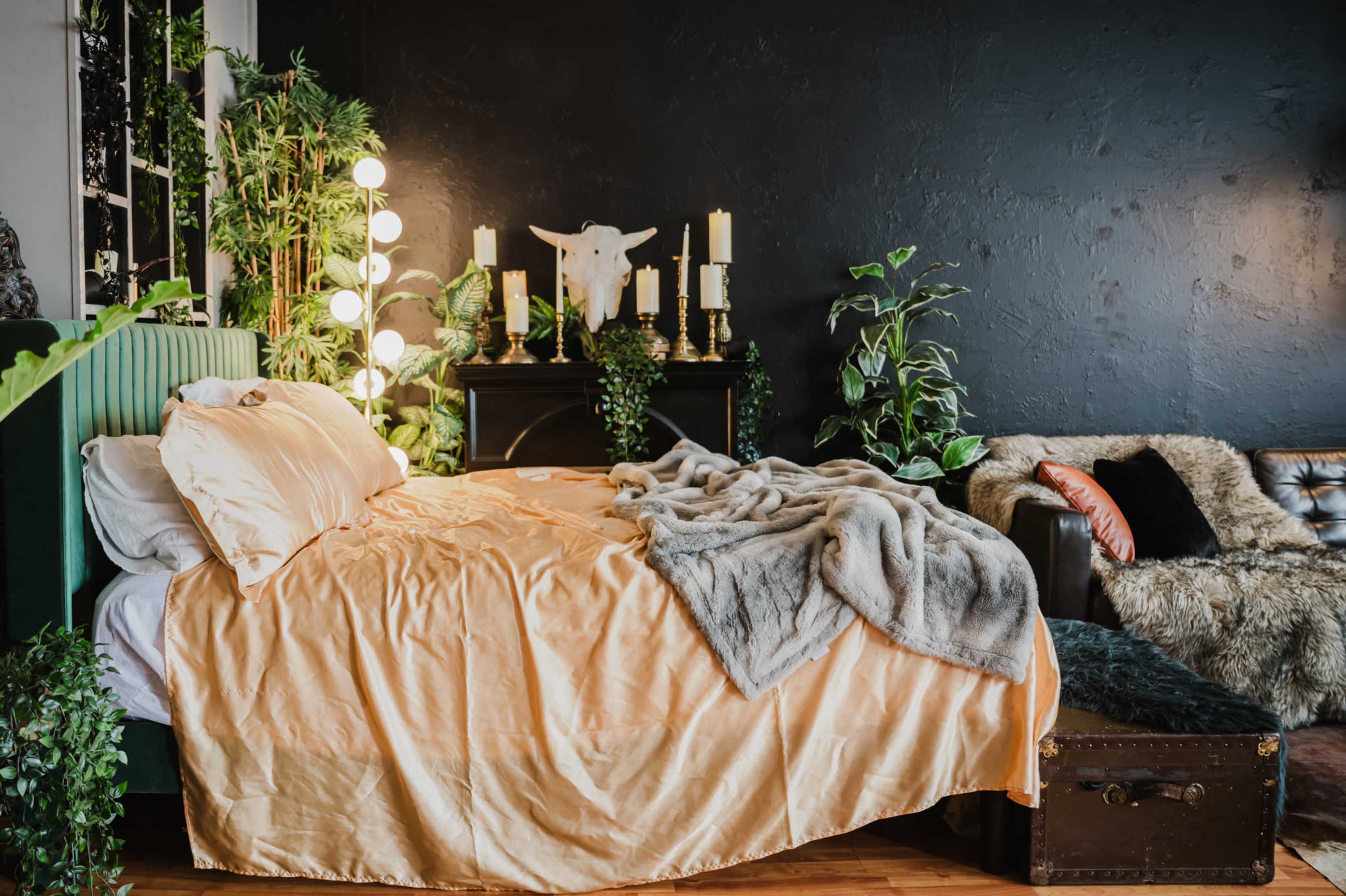 The image shows a cozy bedroom featuring a bed with an orange bedspread, surrounded by plants, candles, and a leather sofa.